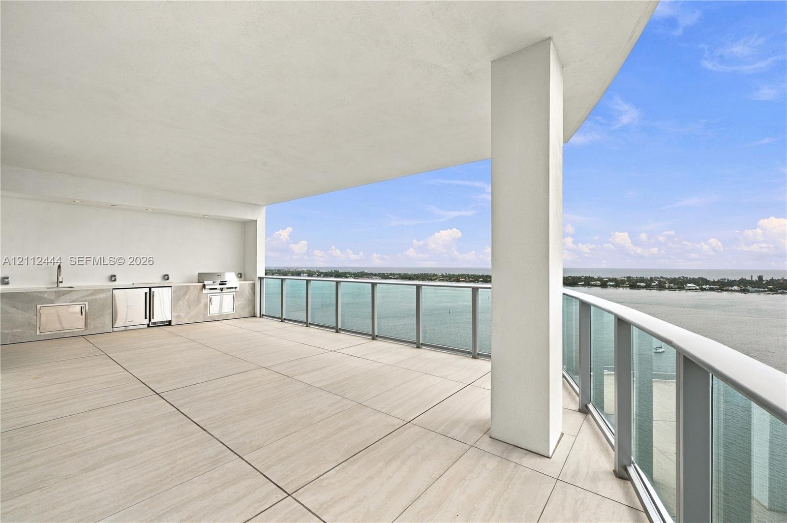 Covered terrace featuring large-format tile flooring, a built-in outdoor kitchen with stone-finish cabinetry, an integrated grill, and a glass-paneled railing