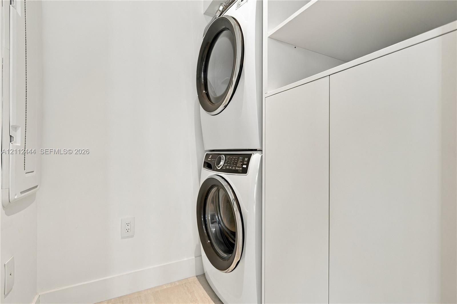 Laundry area featuring a stackable front-load washer and dryer, integrated white cabinetry with open shelving, and light-toned flooring