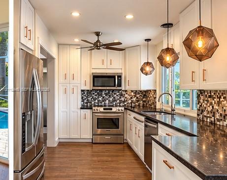 Kitchen featuring white cabinetry with metallic hardware, dark stone-finish countertops, geometric glass pendant lighting, and stainless steel appliances Kitchen featuring white cabinetry with metallic hardware, dark stone-finish countertops, geometric glass pendant lighting, and stainless steel appliances