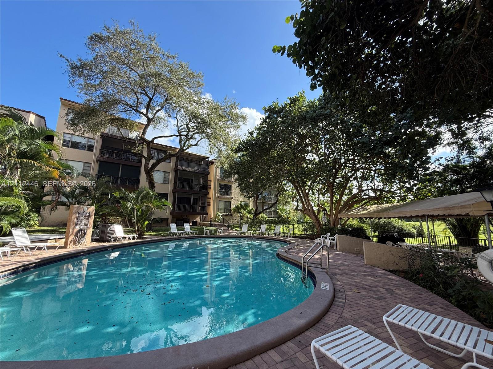 Curved swimming pool featuring a brick paver deck, covered patio area, and mature landscaping
