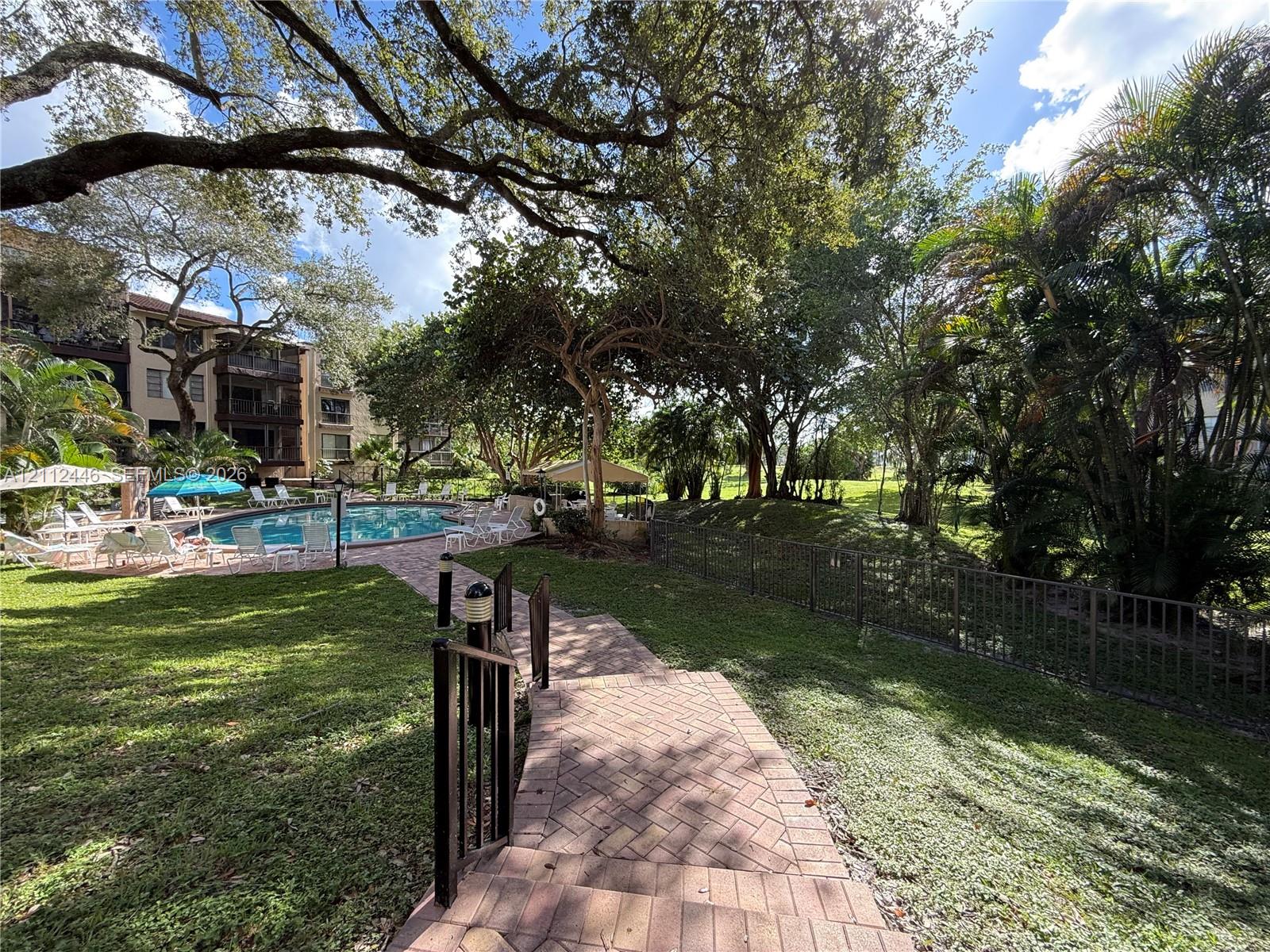 Outdoor pool with brick-paver deck, landscaped grounds, mature shade trees, and metal perimeter fencing