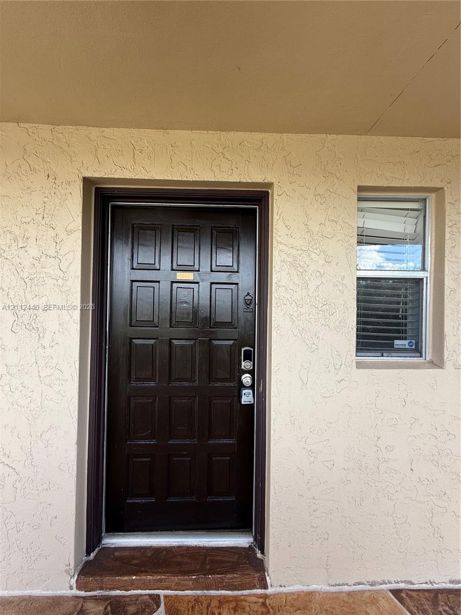 Dark-finished multi-panel entry door with electronic deadbolt, textured stucco exterior, and window with integrated blinds