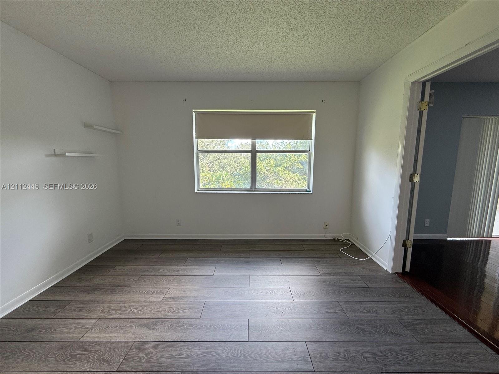 Interior room featuring wood-finish flooring, two wall-mounted shelves, and a single-pane window with a roller shade Interior room featuring wood-finish flooring, two wall-mounted shelves, and a single-pane window with a roller shade