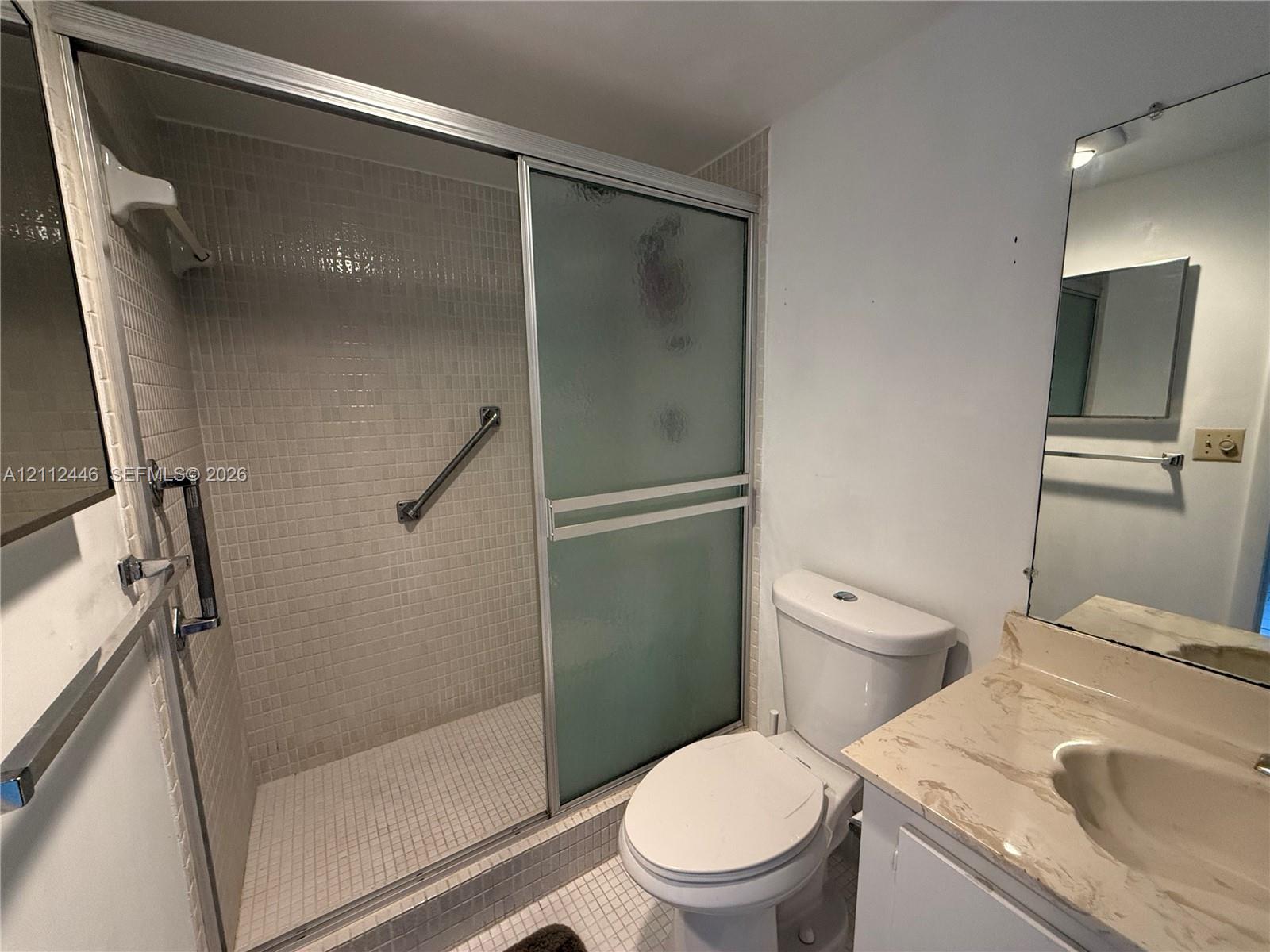 Bathroom featuring a walk-in shower with frosted sliding glass doors, mosaic tile surround, and mounted grab bars, paired with a vanity with a stone-finish countertop and integrated sink Bathroom featuring a walk-in shower with frosted sliding glass doors, mosaic tile surround, and mounted grab bars, paired with a vanity with a stone-finish countertop and integrated sink