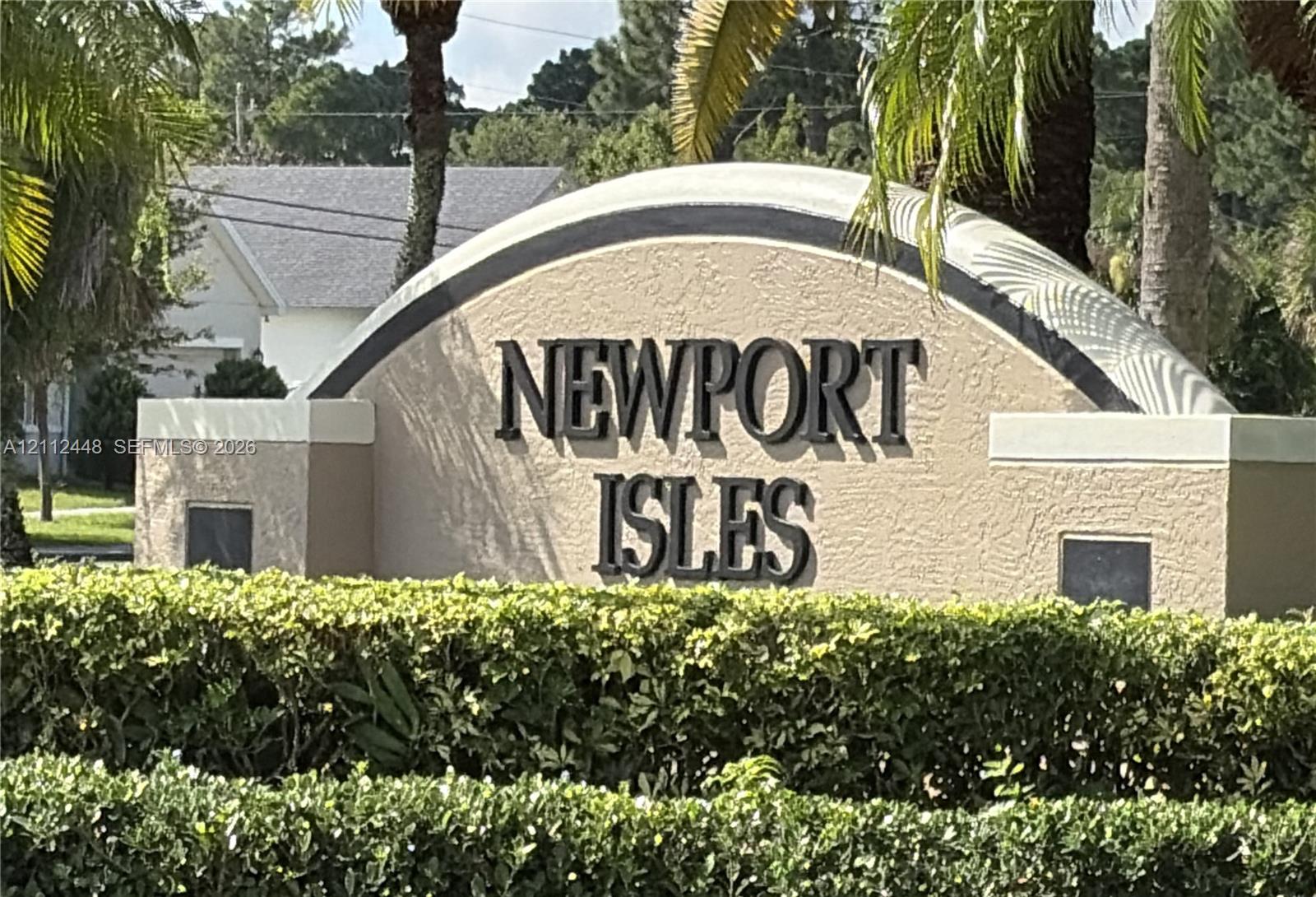 Community monument sign featuring stucco construction, arched detailing, and elevated lettering, accented by mature perimeter hedging and palm trees