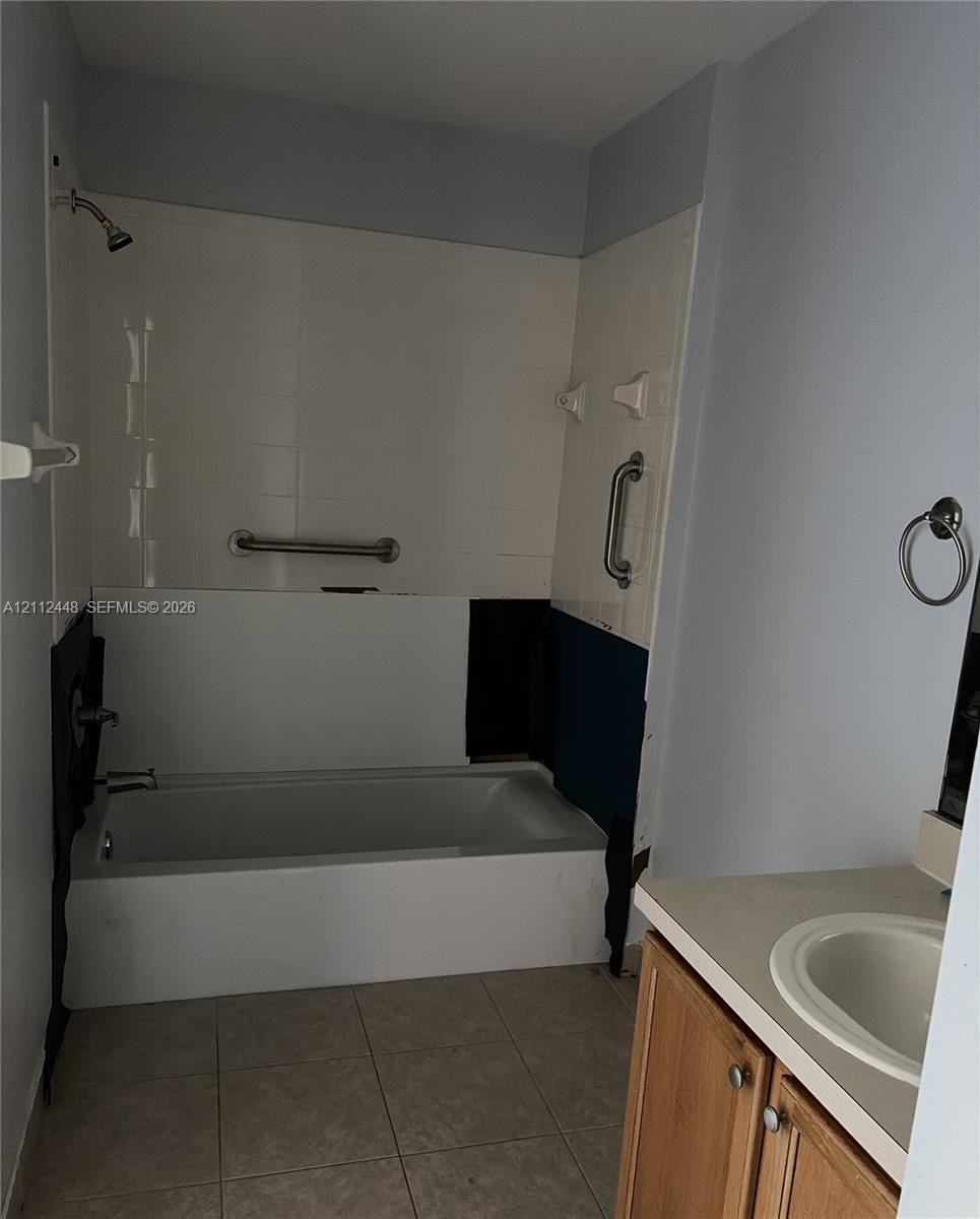 Bathtub with wall-mounted shower head, tile surround, and installed safety grab bars, paired with a wood-finish vanity featuring an integrated basin and a neutral countertop