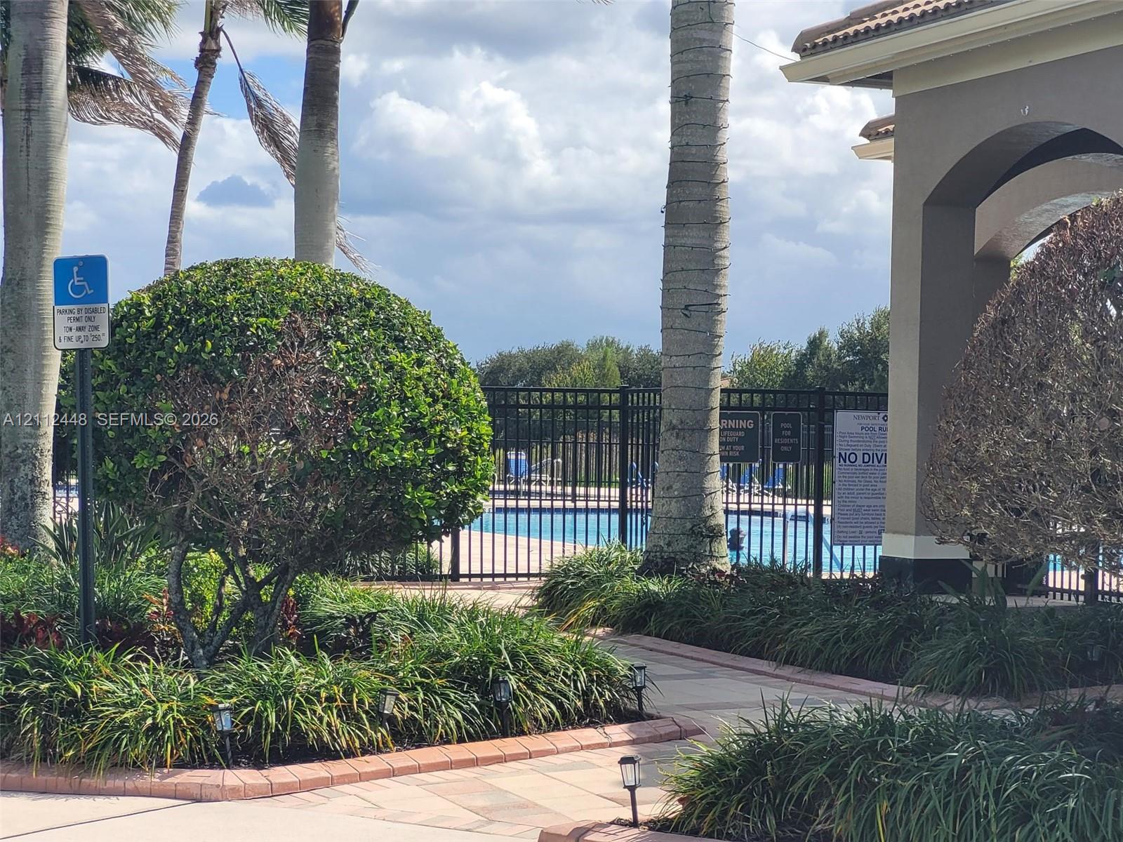 Swimming pool enclosed by a metal fence, bordered by stone-paver walkways, mature palm trees, and manicured landscaping