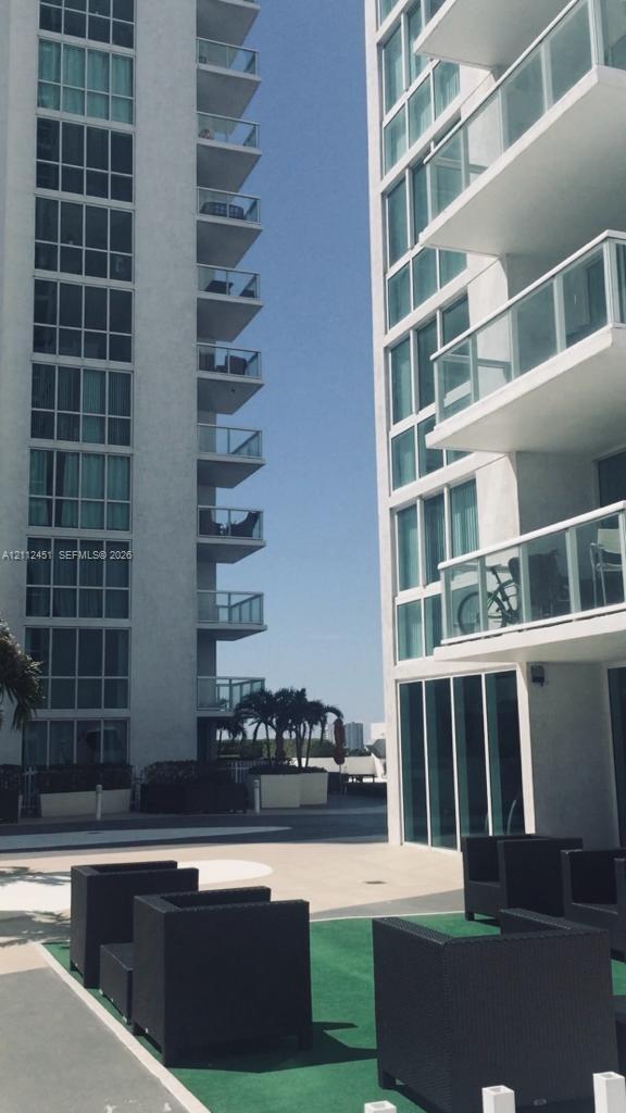 High-rise exterior featuring floor-to-ceiling glass windows, stacked private balconies with glass railings, and a concrete-paved outdoor terrace area