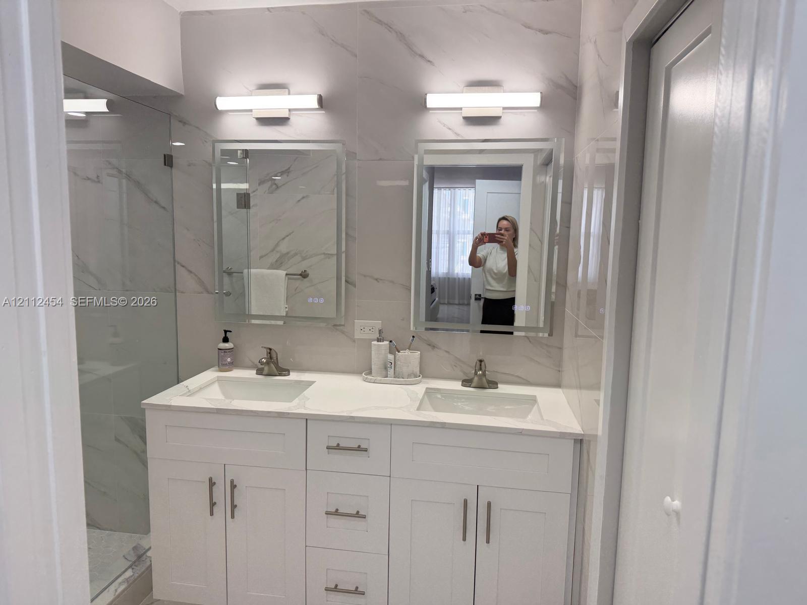 Double vanity featuring white shaker-style cabinetry, white stone-composite countertops, integrated rectangular sinks, and illuminated wall-mounted mirrors with large-format tile surround