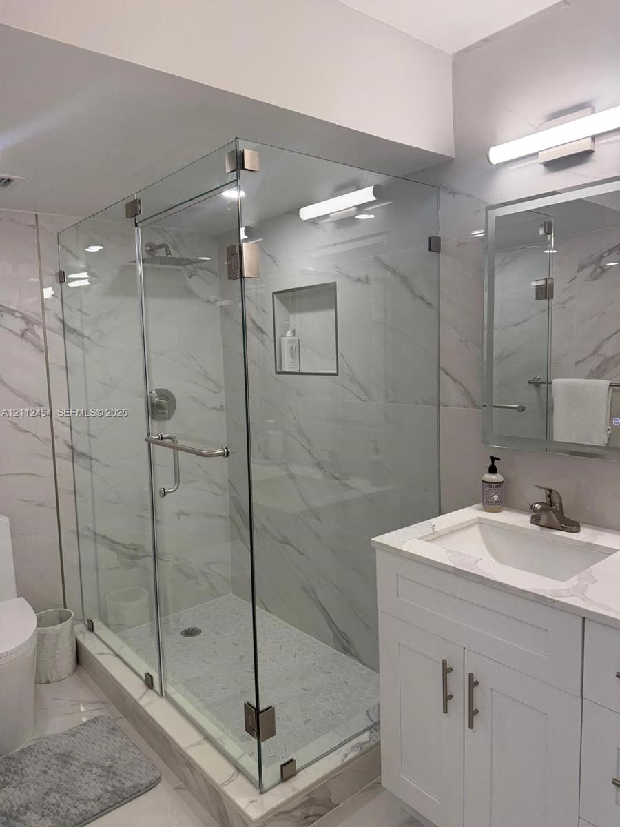 Walk-in shower featuring a frameless glass enclosure, recessed wall niche, and floor-to-ceiling veined stone-finish tile, complemented by a white vanity with a stone-finish countertop and an integrated mirror