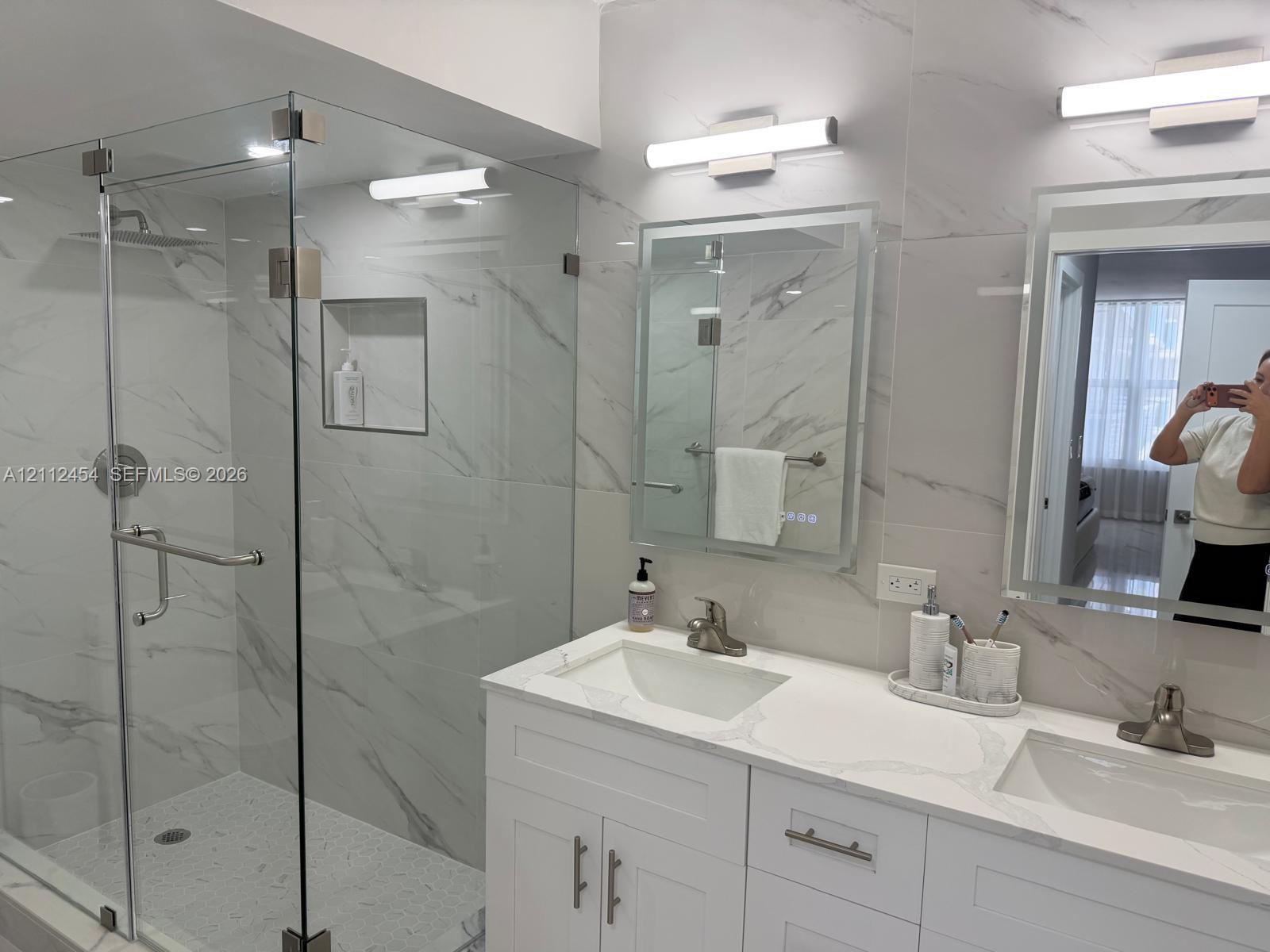 Dual-sink vanity with white cabinetry and stone countertop, glass-enclosed walk-in shower with integrated wall niche, floor-to-ceiling stone-style wall tile, and wall-mounted light fixtures