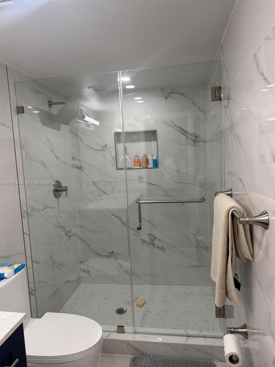 Walk-in shower featuring a frameless glass enclosure, floor-to-ceiling stone-pattern tile, integrated recessed shelving, polished metal hardware, and a pebble-style basin floor