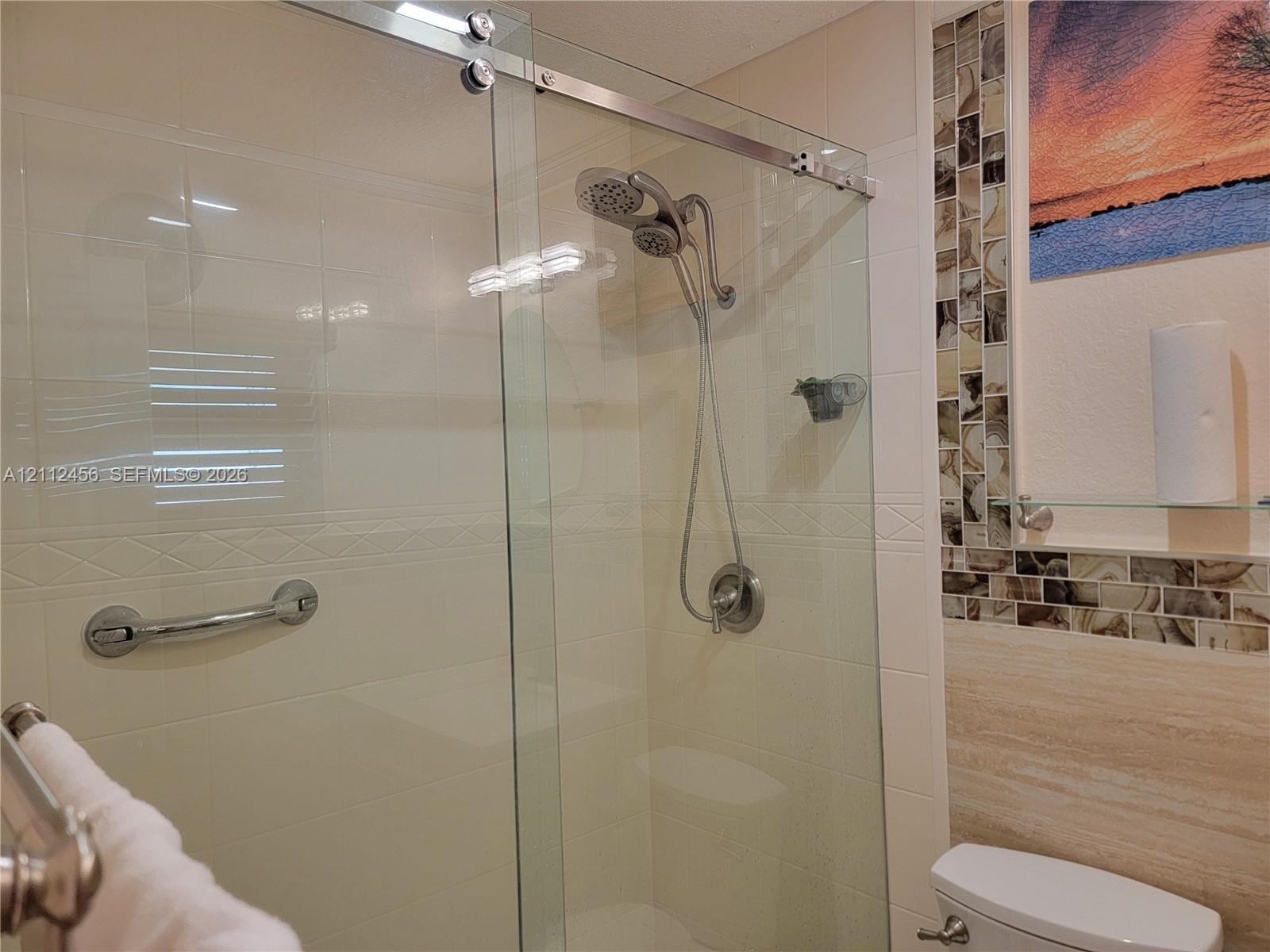 Shower stall featuring a glass sliding door, metallic handheld shower fixture, integrated grab bar, and neutral wall tiling with decorative mosaic accent strips