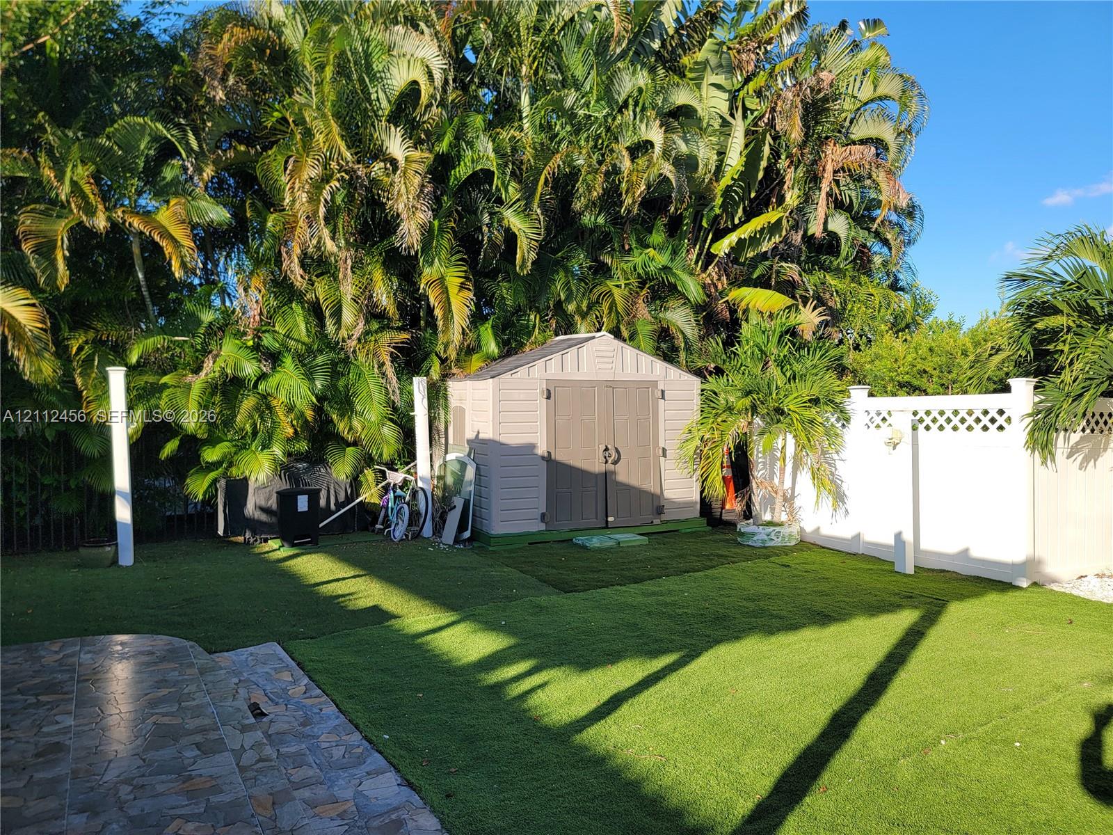 Rear exterior featuring synthetic turf lawn, white vinyl privacy fencing with lattice detail, stone-paver patio, and exterior storage shed