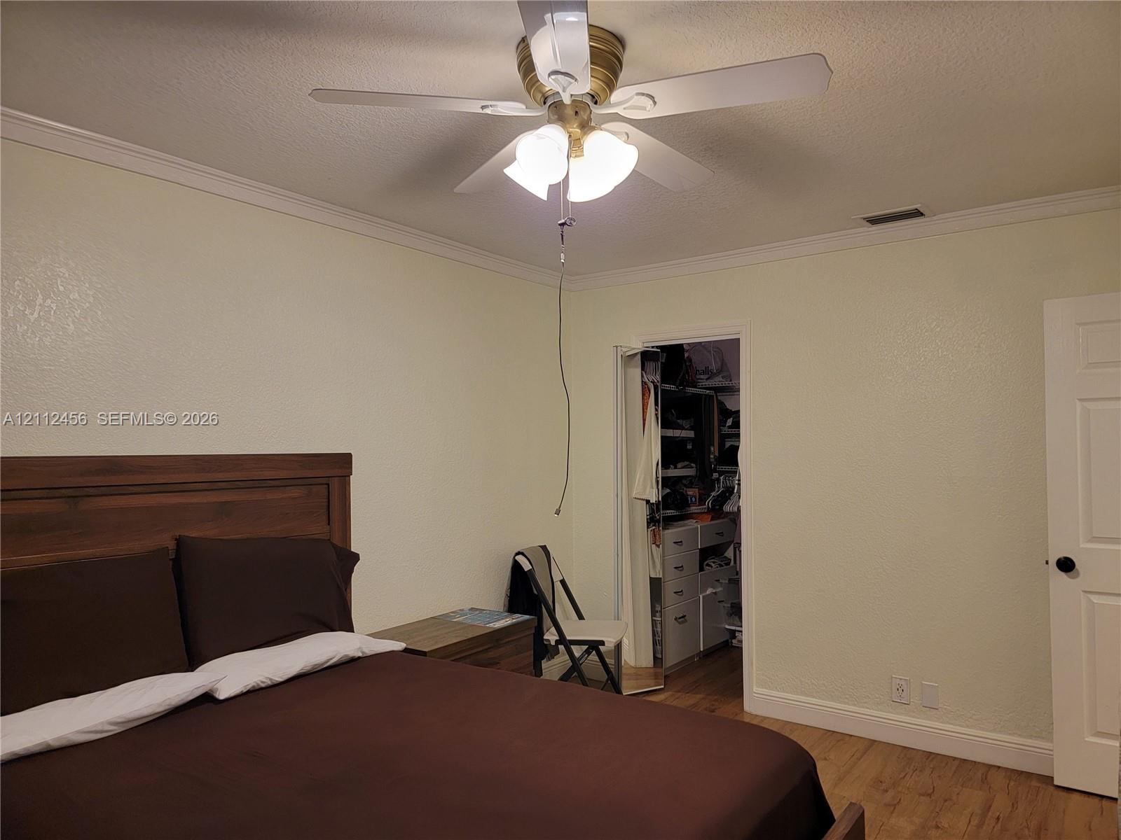 Neutral-toned interior featuring wood-finish flooring, crown molding, a central ceiling fan with multi-light fixture, and an open walk-in closet