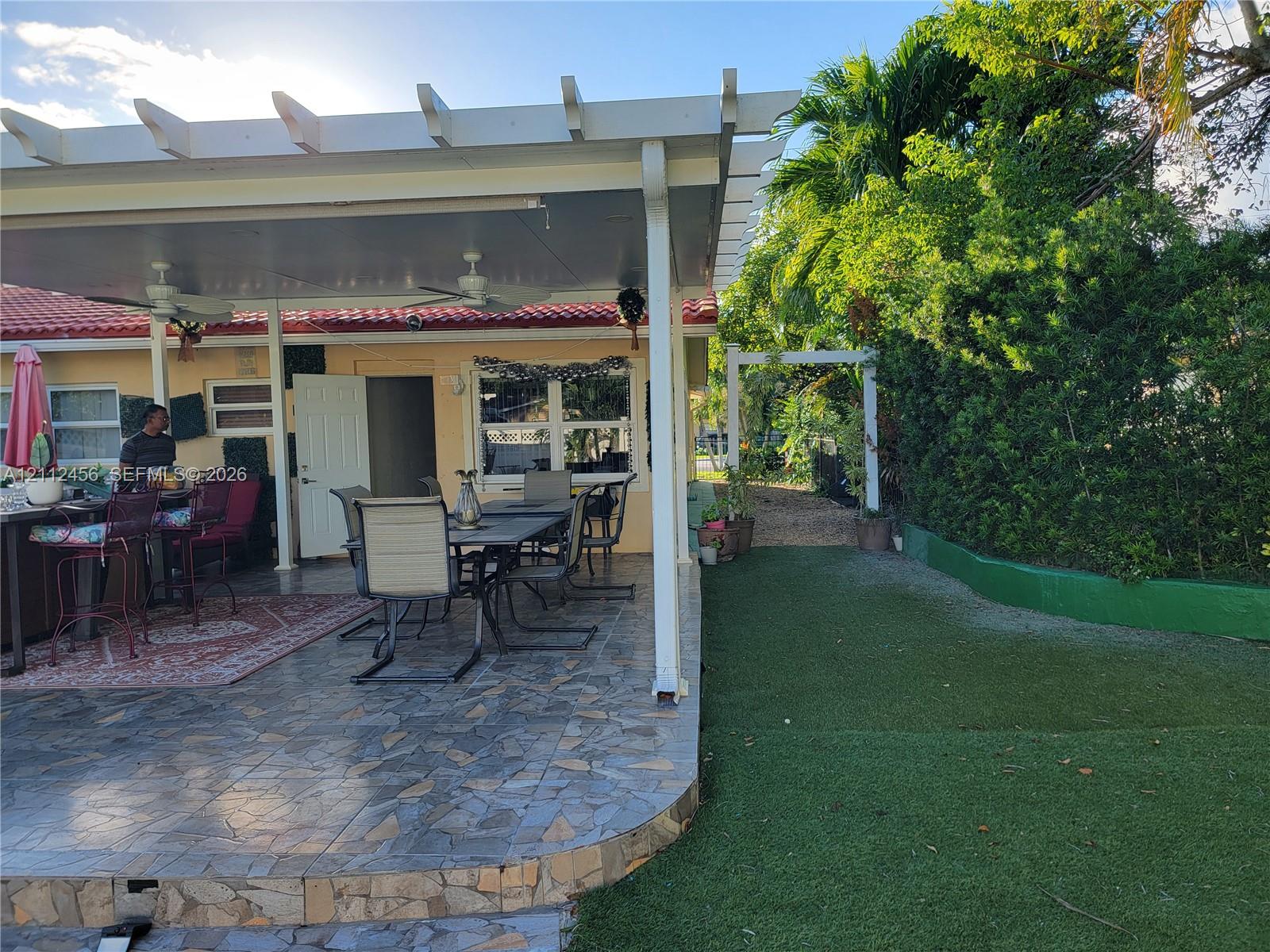 Covered patio with a pergola extension, multi-tone tiled flooring, artificial turf lawn, and mature perimeter landscaping