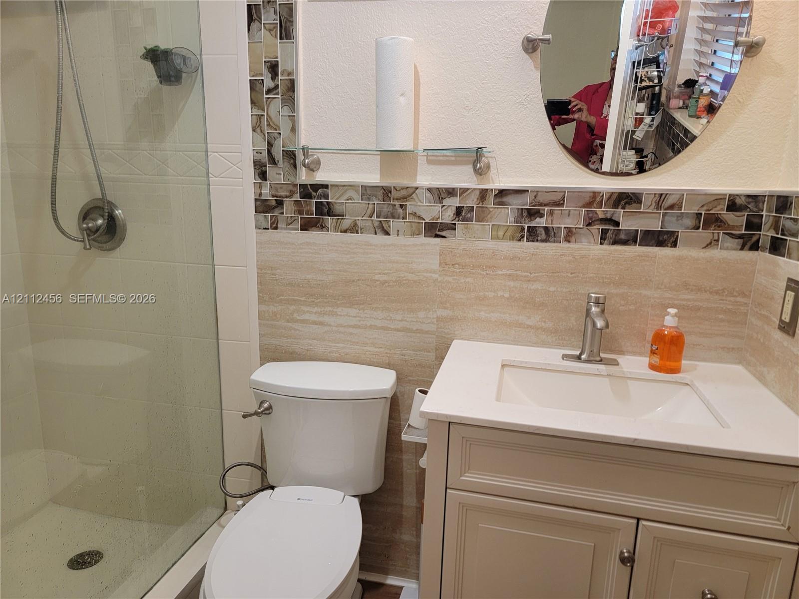 Bathroom featuring a vanity with a white countertop and rectangular undermount sink, stone-finish wall tiling, decorative mosaic tile accents, a glass-enclosed shower, and an oval wall-mounted mirror