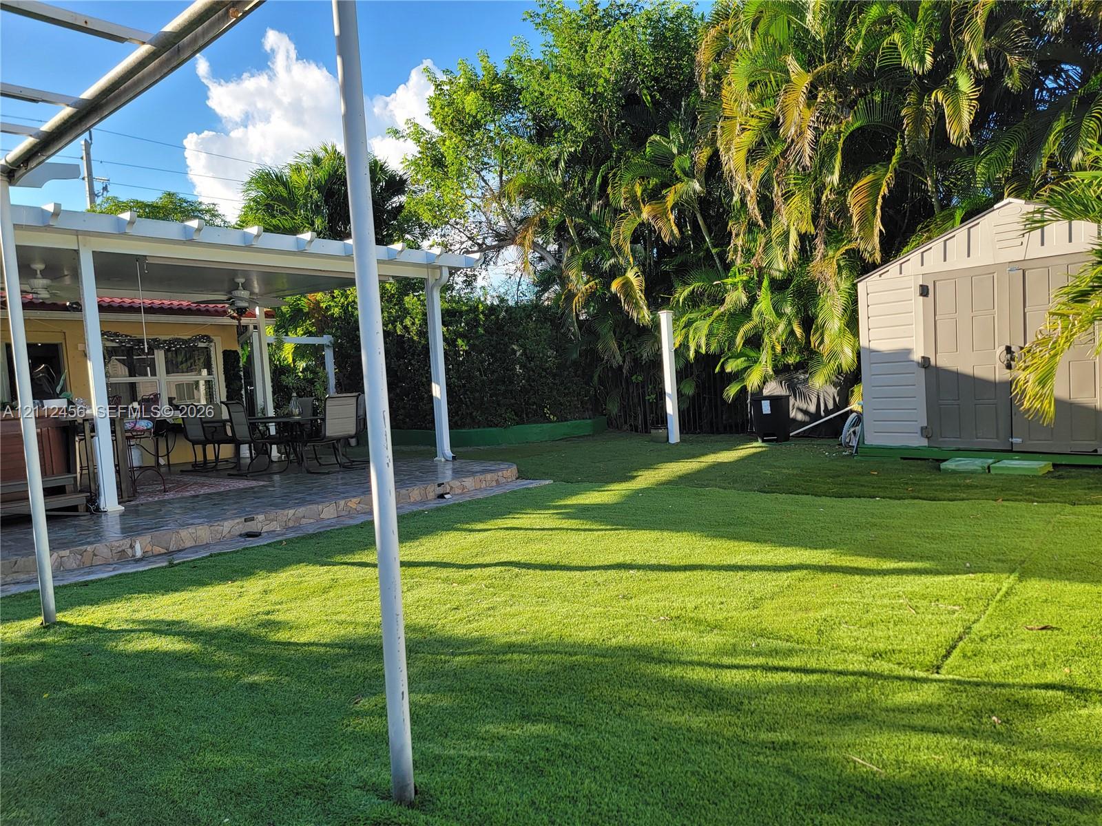 Rear patio featuring a covered pergola, tiled flooring, artificial turf lawn, and a structural storage shed