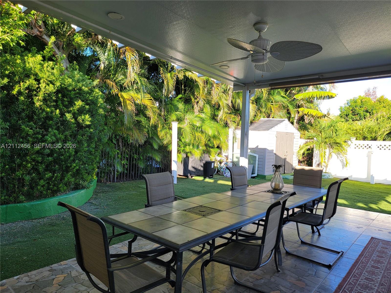 Covered rear patio featuring a tile-paved floor and ceiling-mounted fan, overlooking a lawn with mature landscaping and a detached storage shed