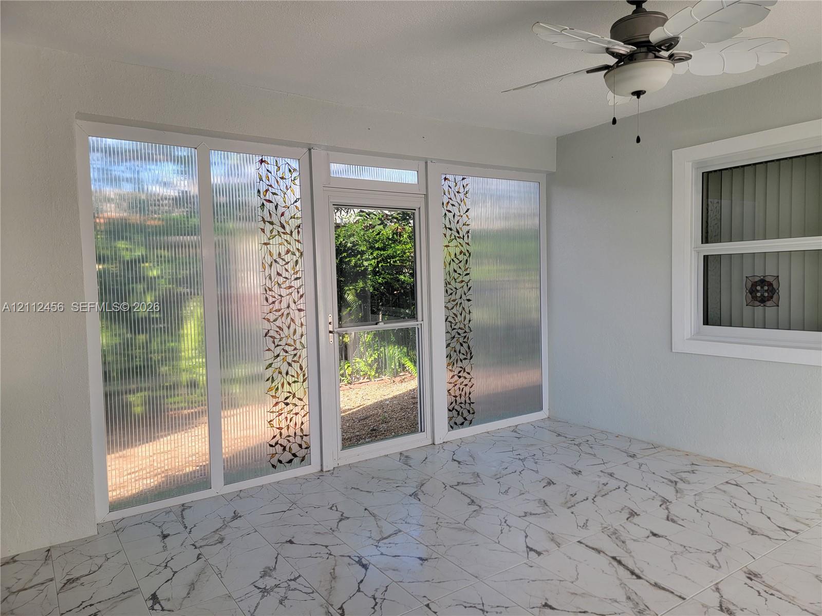 Enclosed space featuring large format stone-patterned tile flooring, textured glass panels with decorative leaf detailing, a central full-view storm door, and a ceiling-mounted fan with integrated light fixture
