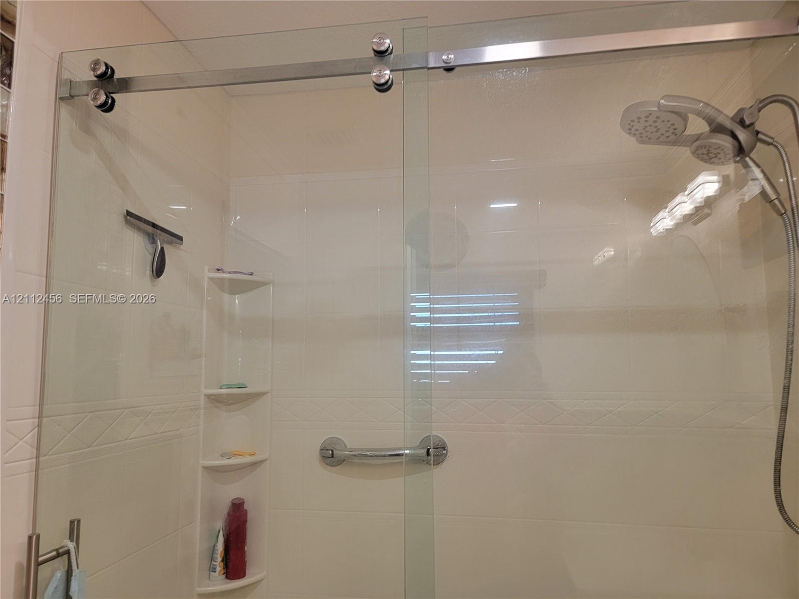Frameless glass shower enclosure featuring a stainless steel sliding door system, integrated corner shelving, chrome grab bar, and adjustable handheld shower head