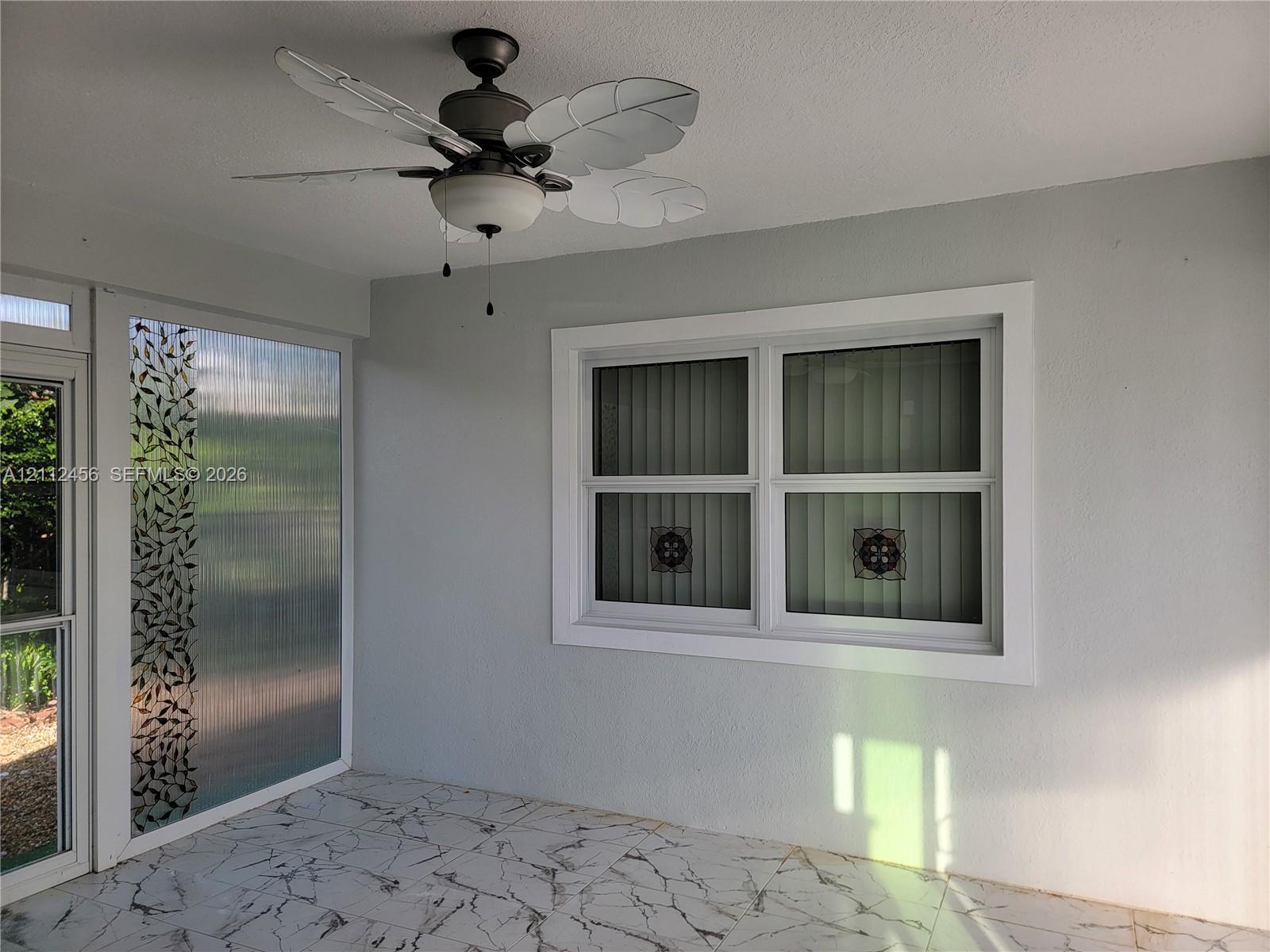 Enclosed patio featuring patterned tile flooring, a dual-light ceiling fan with leaf-shaped blades, and decorative glass paneling