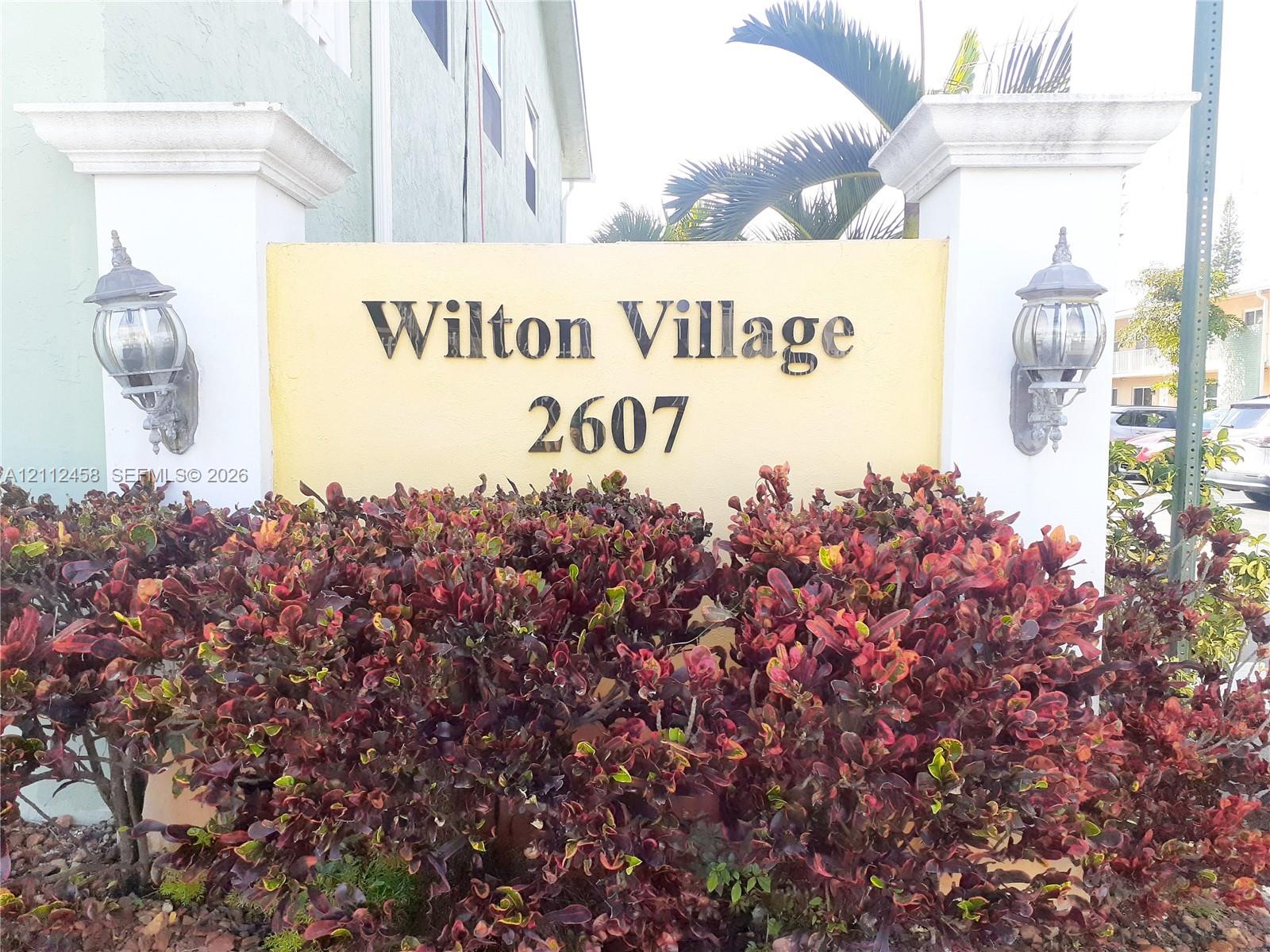 Entrance monument featuring stucco pillars, wall-mounted coach-style lighting, and mature landscape shrubbery