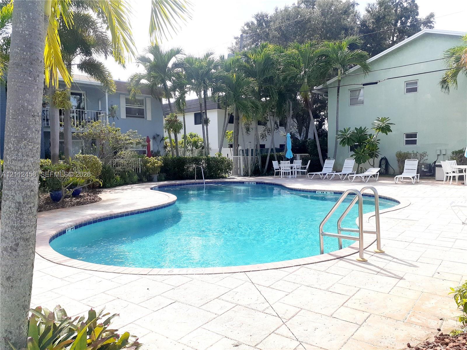 Curved swimming pool with a stone paver deck, metal ladder, and mature palm tree landscaping