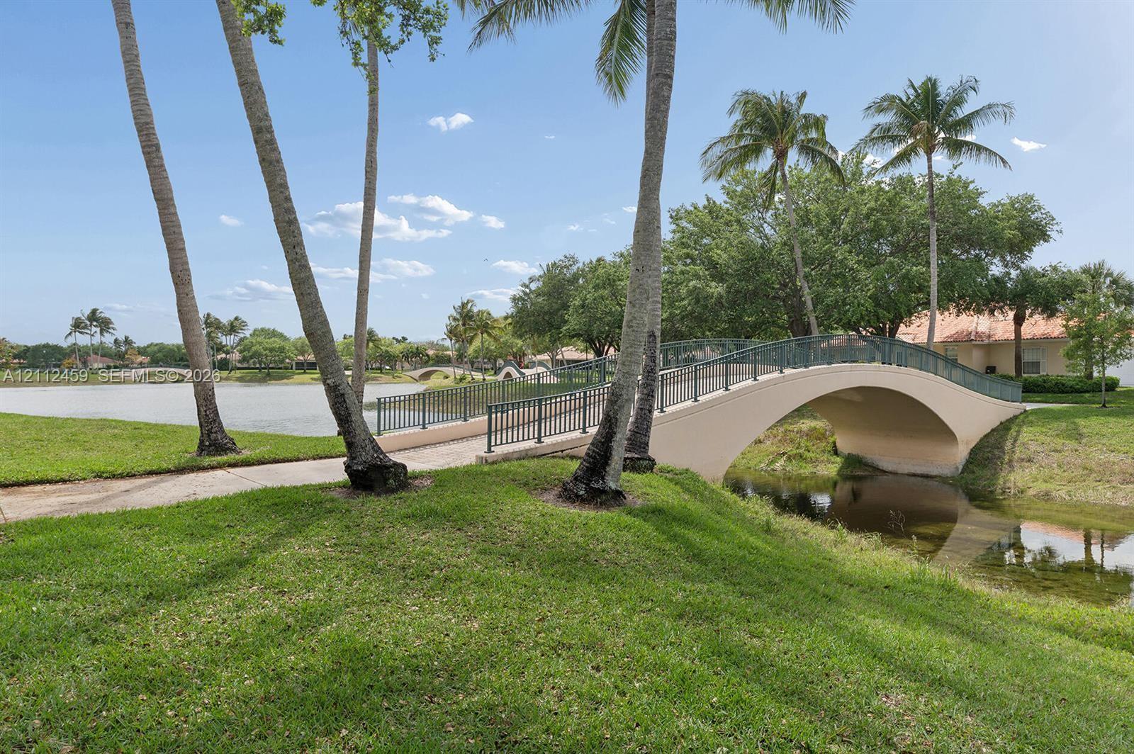 Arched concrete pedestrian bridge featuring metal railings, surrounded by mature palm trees, manicured lawn, and a waterfront setting