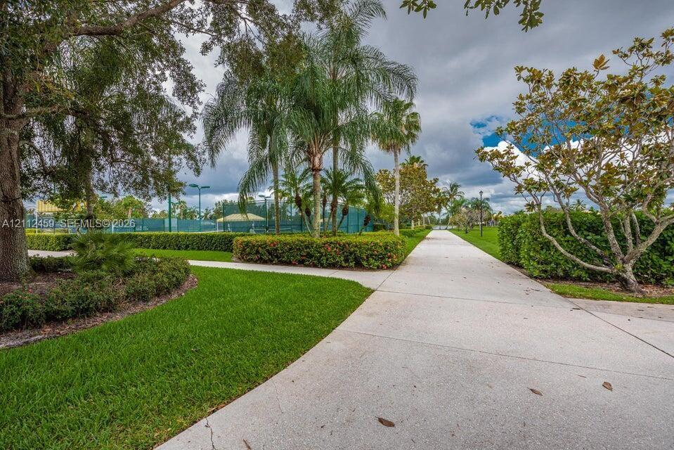 Concrete pedestrian walkway flanked by manicured green space, mature palm trees, sculpted landscaping, and fenced outdoor recreational courts