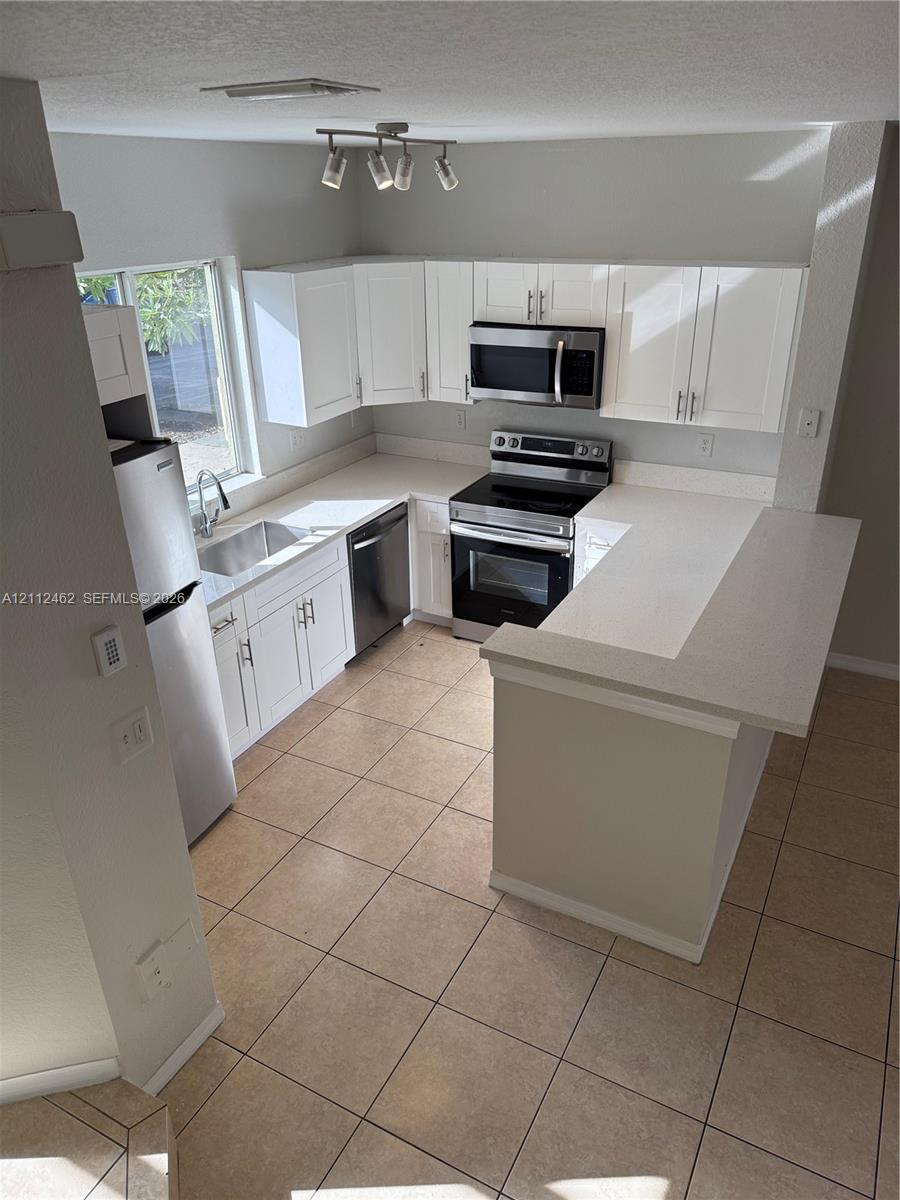 Kitchen featuring white shaker-style cabinetry, solid-surface countertops, tile flooring, stainless steel appliances, and track lighting