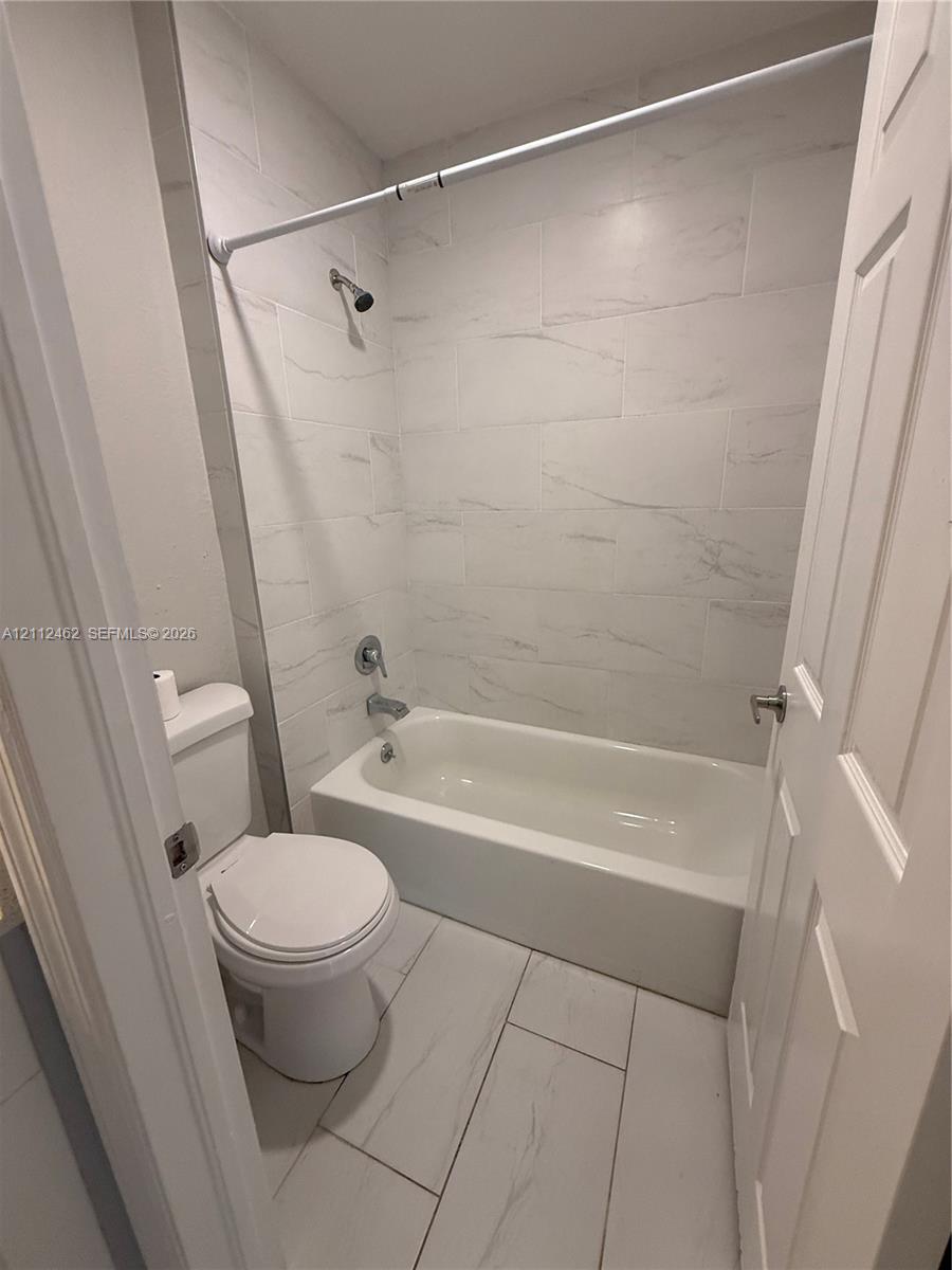 Full bathroom featuring large-format light-toned tile flooring, a tub-shower combination, and wall-to-wall light-toned tile surround