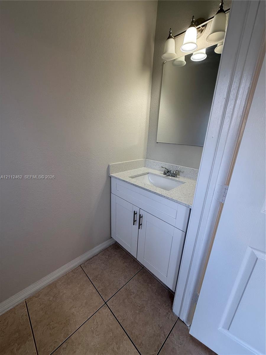 Bathroom vanity featuring white shaker-style cabinetry, solid-surface countertop with an integrated basin, three-light fixture, and ceramic tile flooring