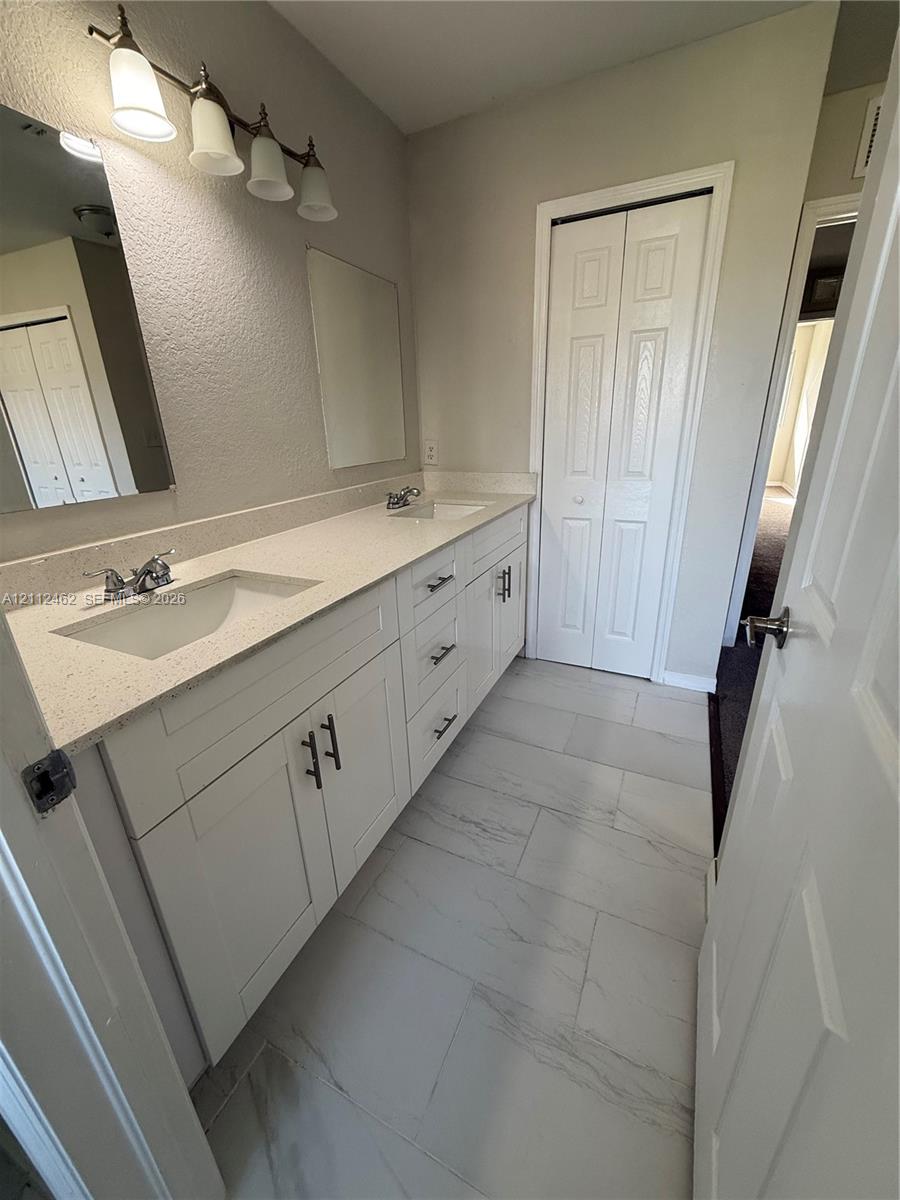 Dual vanity featuring a light-toned stone-composite countertop, white shaker-style cabinetry with matte black hardware, integrated rectangular basins, and large format rectified tile flooring
