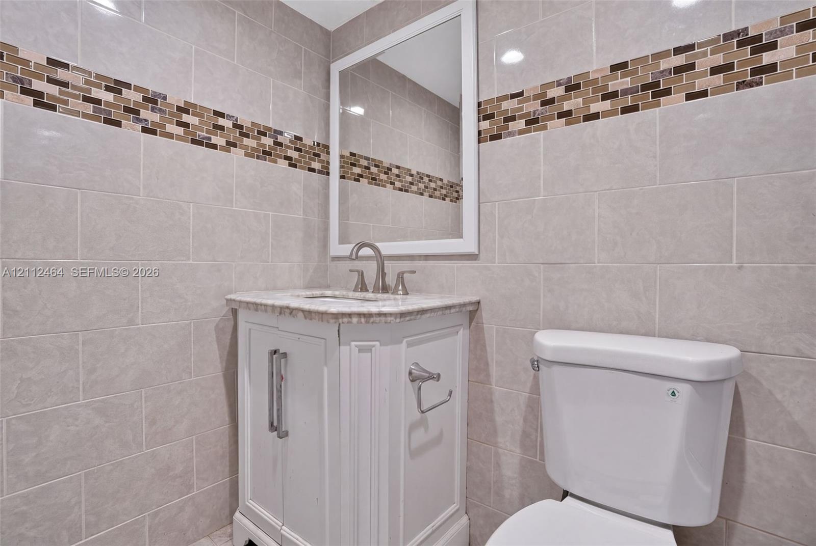 Wall-mounted mirror with white frame, vanity with stone-finish countertop, brushed nickel faucet, and neutral wall tile with a mosaic accent border