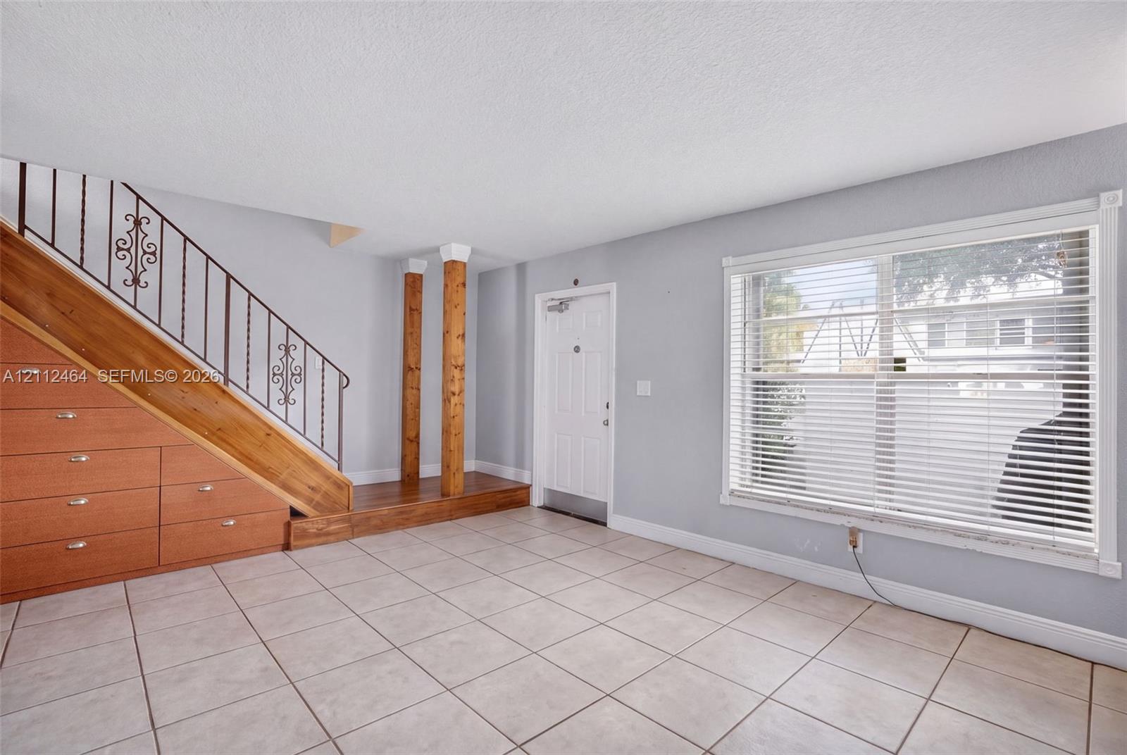 Interior featuring tile flooring, a large window with horizontal blinds, built-in wooden under-stair storage, decorative wrought-iron railing, and dual wooden support columns Interior featuring tile flooring, a large window with horizontal blinds, built-in wooden under-stair storage, decorative wrought-iron railing, and dual wooden support columns