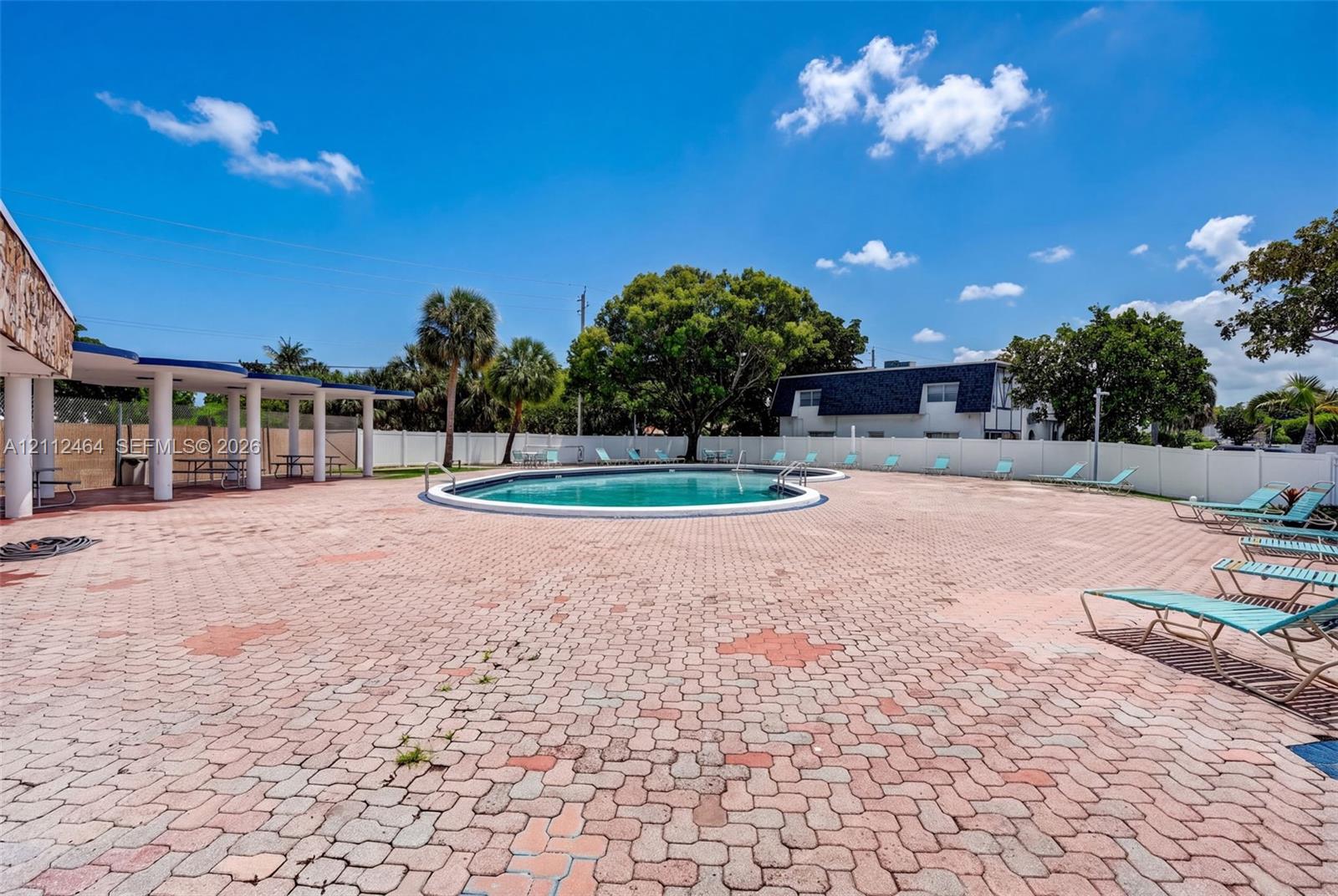 Swimming pool with a circular basin, surrounded by a paved brick patio, covered pavilion area with structural columns, and perimeter privacy fencing