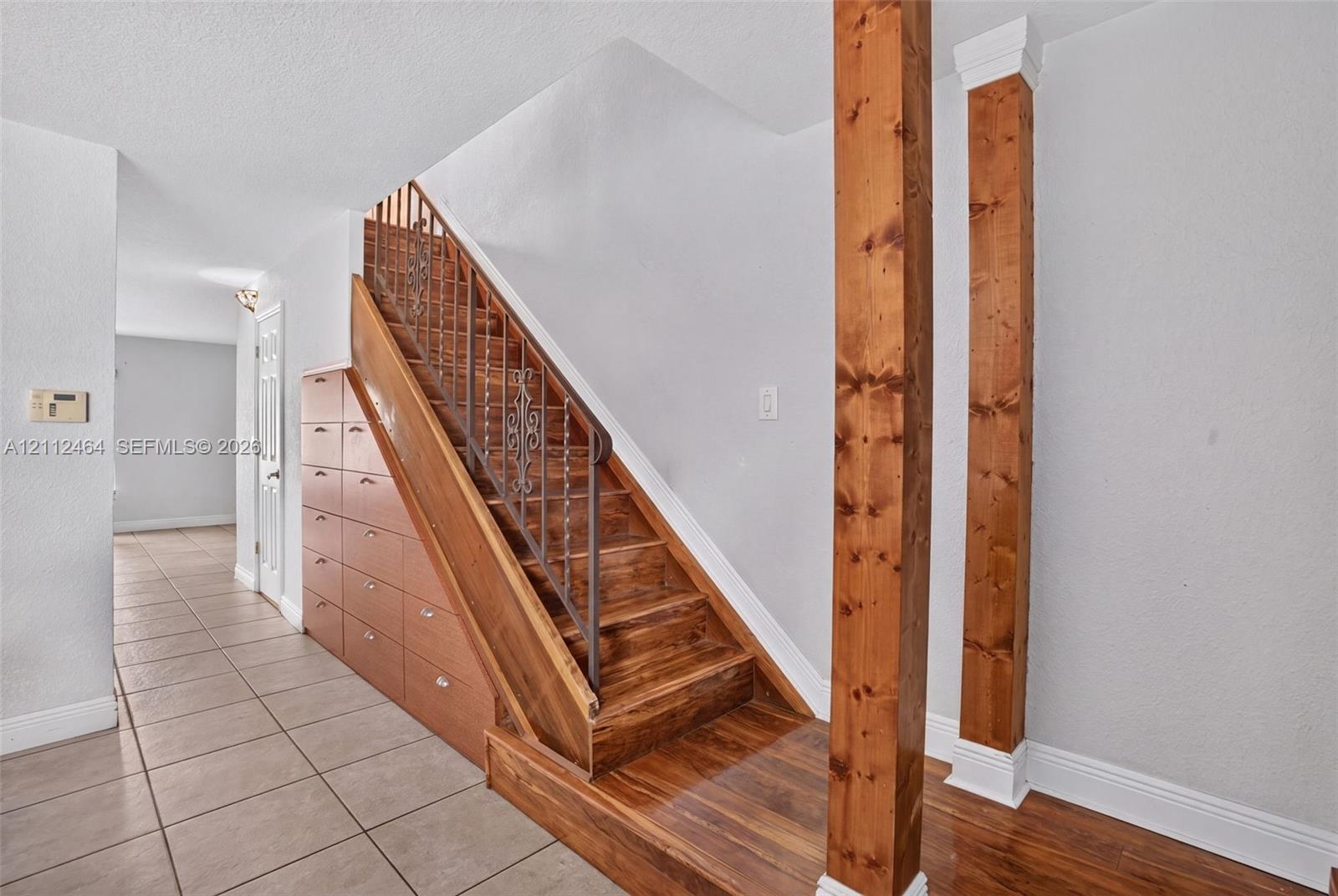 Staircase featuring wood treads, ornate metal handrail, and integrated under-stair cabinetry, accented by two prominent wood-finish structural columns Staircase featuring wood treads, ornate metal handrail, and integrated under-stair cabinetry, accented by two prominent wood-finish structural columns