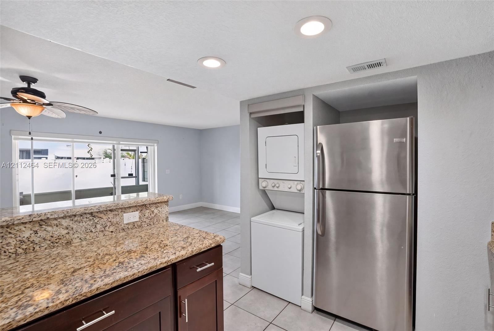 Kitchen area featuring stone-finish countertops, dark wood-finish cabinetry, stainless steel refrigerator, stacked laundry unit, and ceramic tile flooring