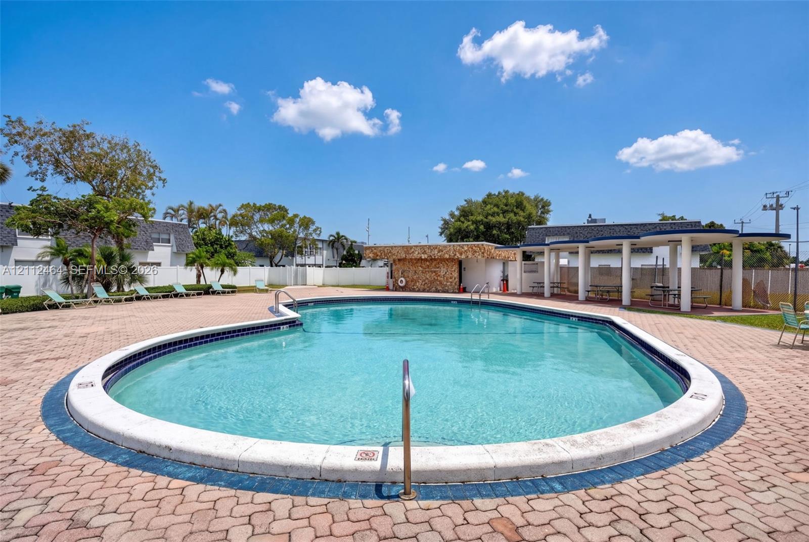 Curved swimming pool with a stone-accented pool house, expansive paved patio, and a covered pergola area