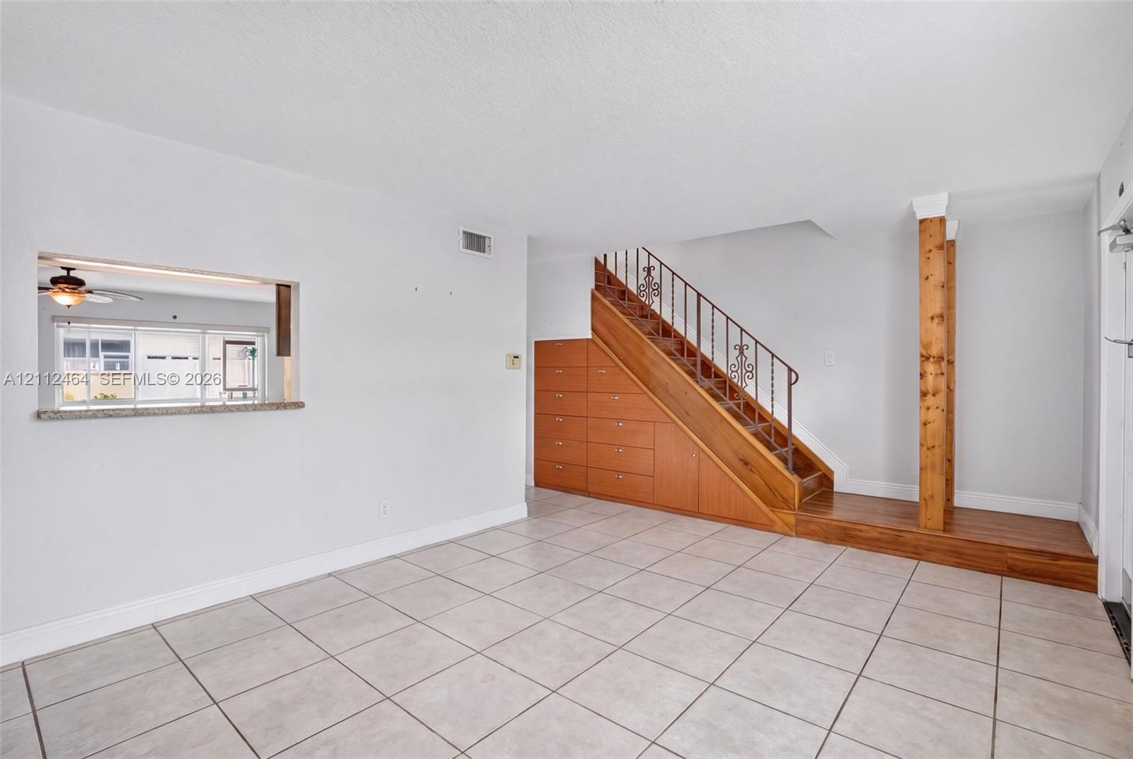 Interior space featuring large-format tile flooring, built-in under-stair cabinetry with wood finish, decorative metal stair railing, and a pass-through window with a stone-finish ledge Interior space featuring large-format tile flooring, built-in under-stair cabinetry with wood finish, decorative metal stair railing, and a pass-through window with a stone-finish ledge