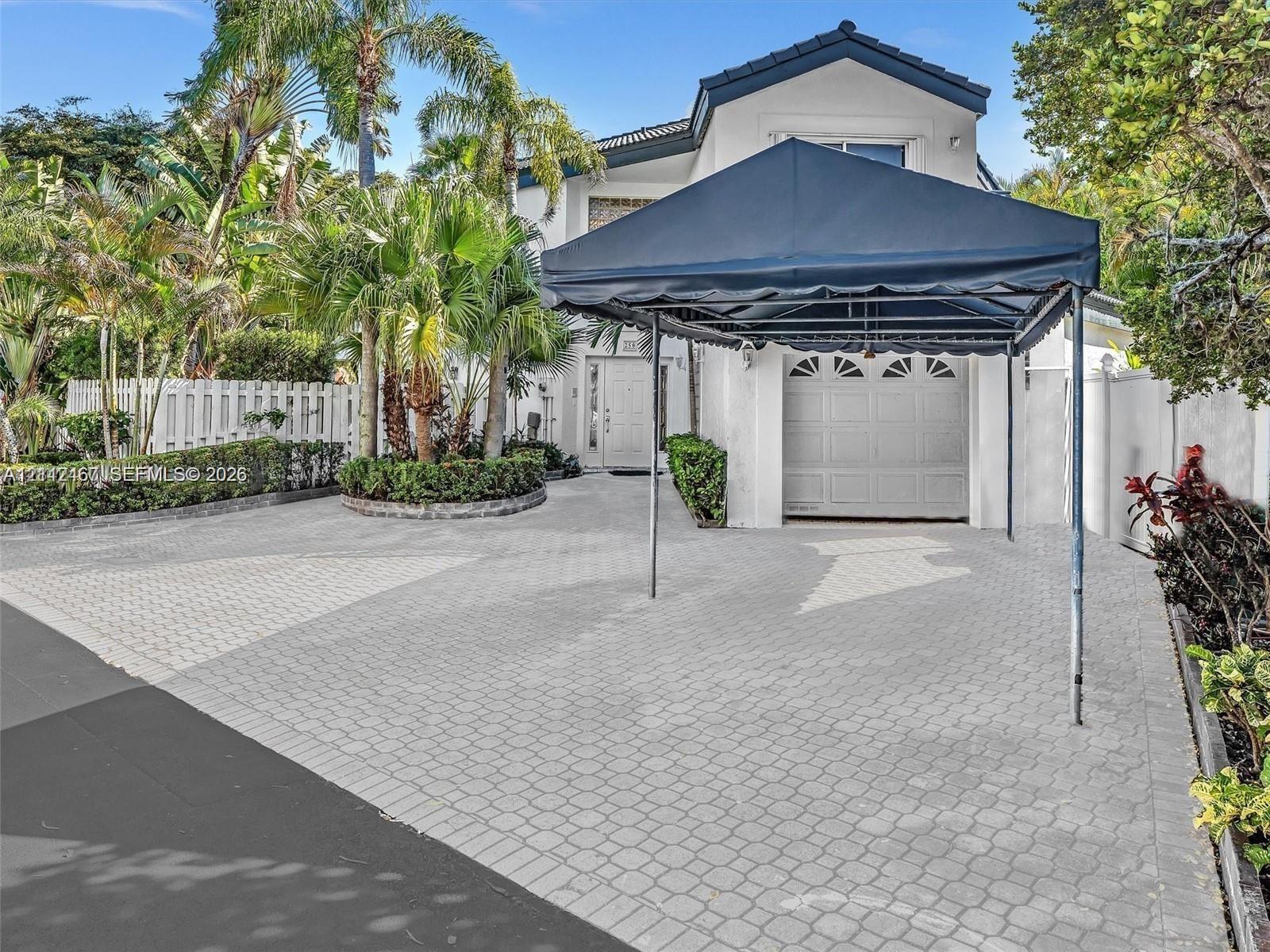 Exterior featuring a paved driveway, mature palm trees, multi-paneled garage door, and white privacy fencing