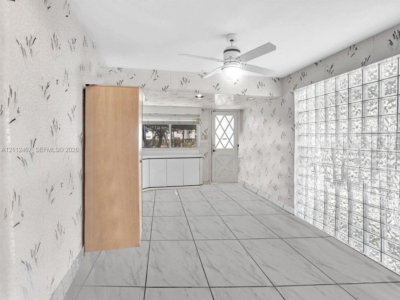 Room featuring large-format stone-patterned tile flooring, a floor-to-ceiling glass block wall, built-in light wood cabinetry, a white cross-panel door, and a ceiling-mounted light fixture with fan blades