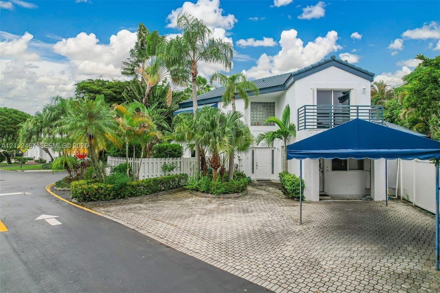 Two-story exterior featuring a paver driveway, covered carport, private balcony with metal railing, and mature palm landscaping