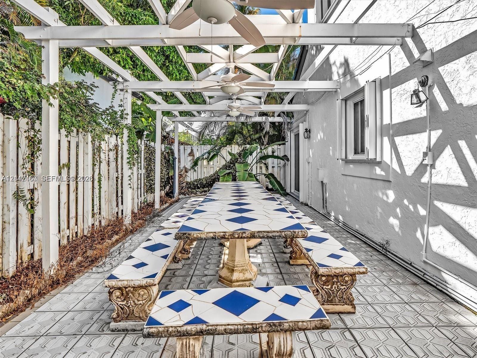 Outdoor patio featuring a white pergola with ceiling-mounted fans, geometric tile flooring, built-in tile-topped dining surfaces, and perimeter wood fencing
