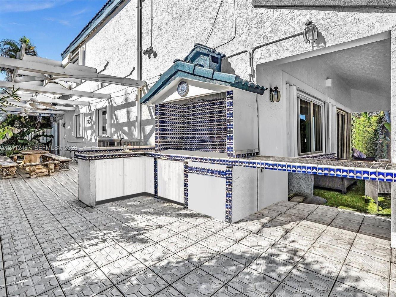 Outdoor kitchen featuring a built-in grill, tiled countertop, decorative patterned backsplash, and a gabled roof structure, complemented by a expansive tiled patio and a white wooden pergola