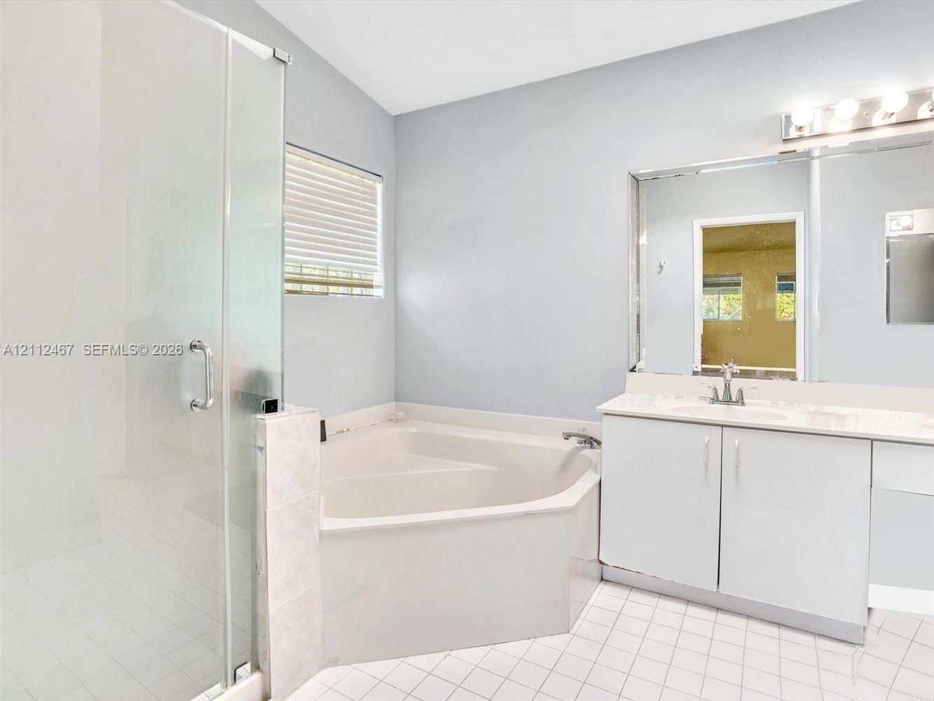 Glass-enclosed shower with tiled flooring, corner soaking tub, vanity with stone countertop, oversized wall-mounted mirror, and multi-bulb vanity lighting