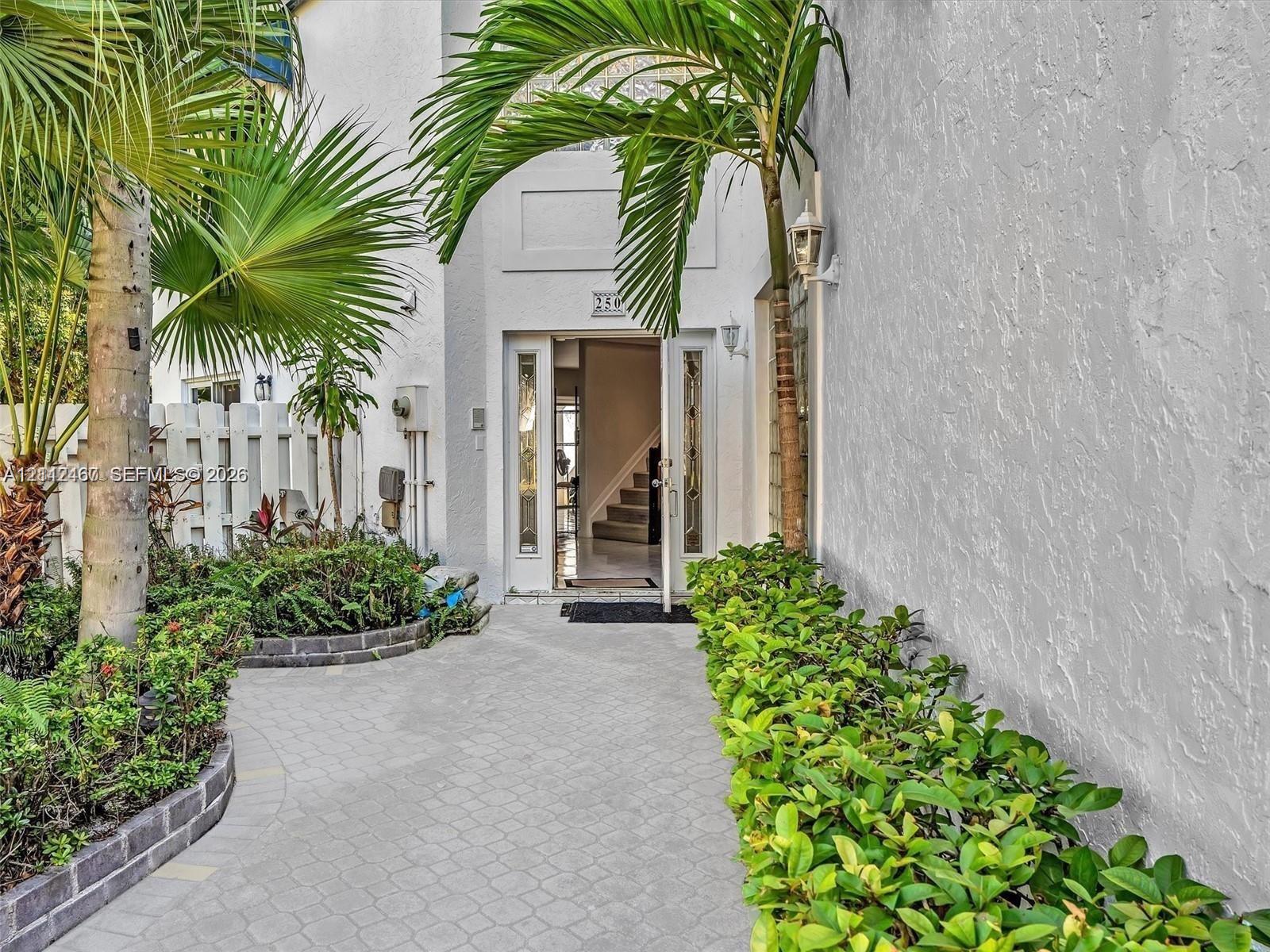 Stucco exterior entrance featuring a paver walkway, mature palm trees, decorative landscaping, and glass-panel door inserts