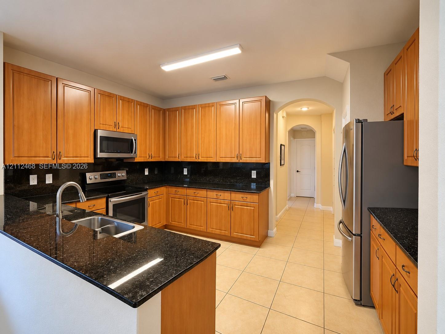 Kitchen featuring wood cabinetry, dark stone countertops, stainless steel appliances, tile flooring, and an arched doorway