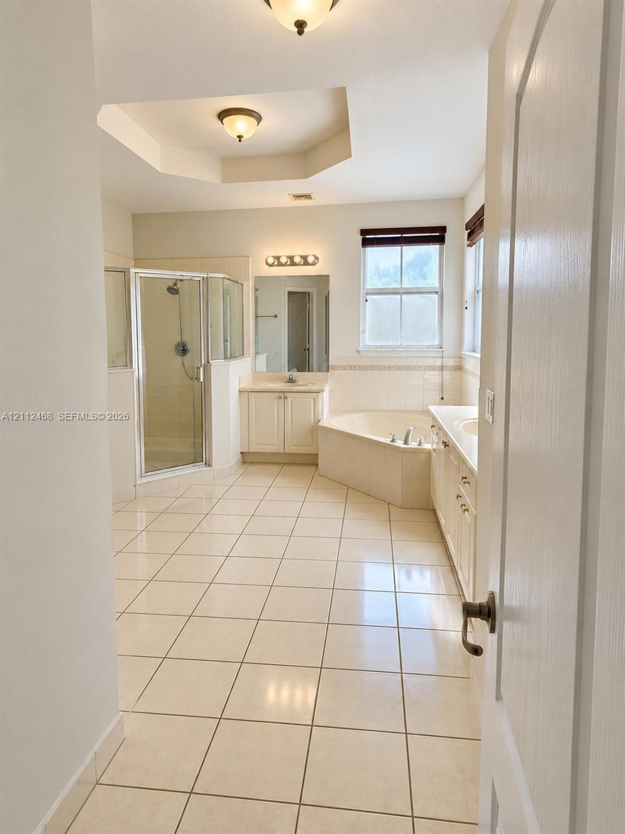 Ceramic tile flooring, corner soaking tub with tile surround, glass-enclosed shower, dual-sink vanity with light-colored cabinetry, and recessed tray ceiling